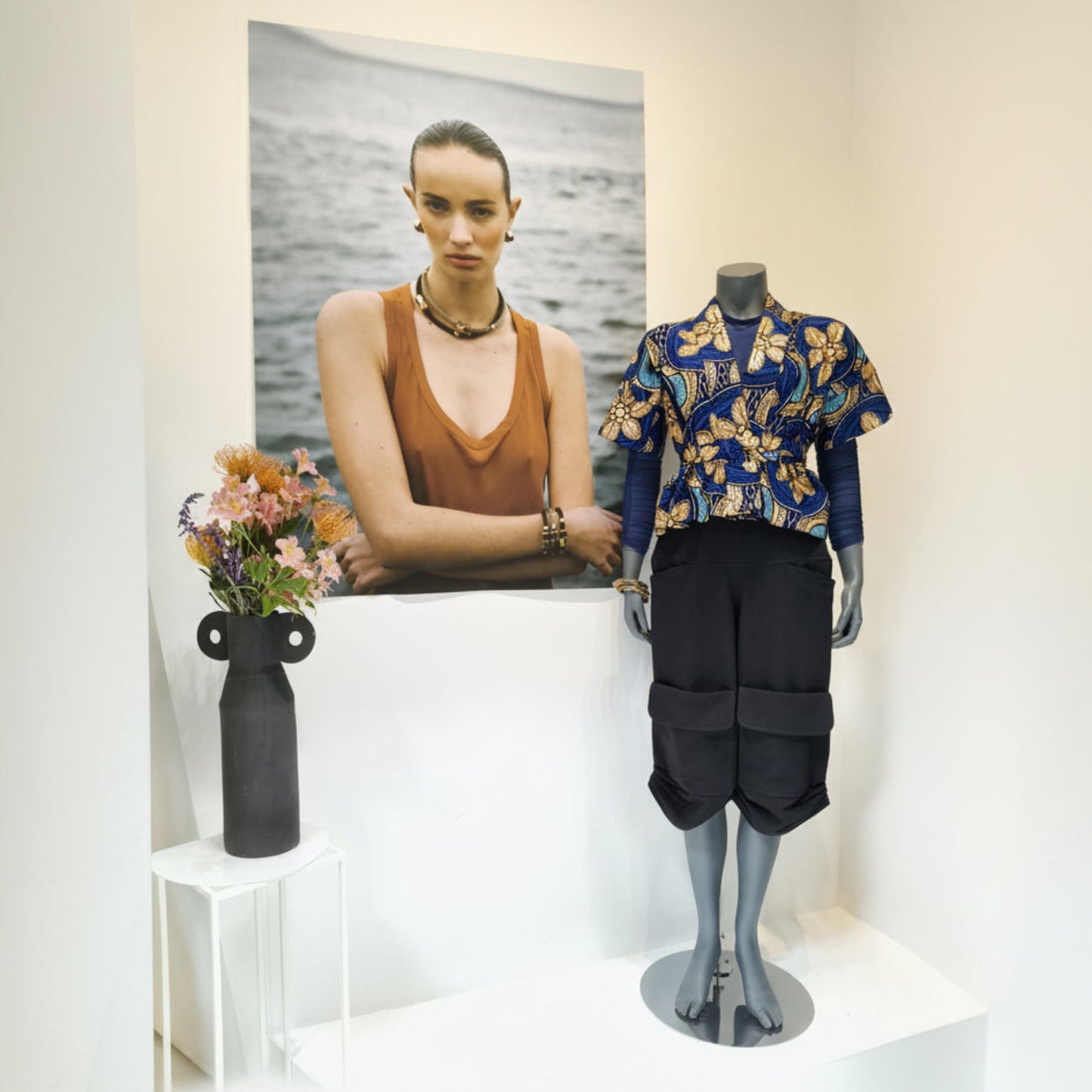 A mannequin displays a blue short-sleeve button-up shirt with a yellow and white floral print, paired with black cargo shorts. Next to it is a white side table holding a black vase with flowers, and behind the mannequin is a large framed photograph of a woman.
