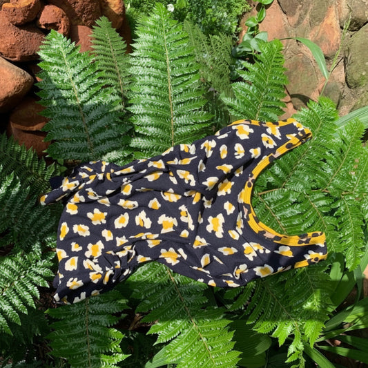 A black sleeveless top with a yellow and white floral pattern, laid flat on a bed of green fern leaves.