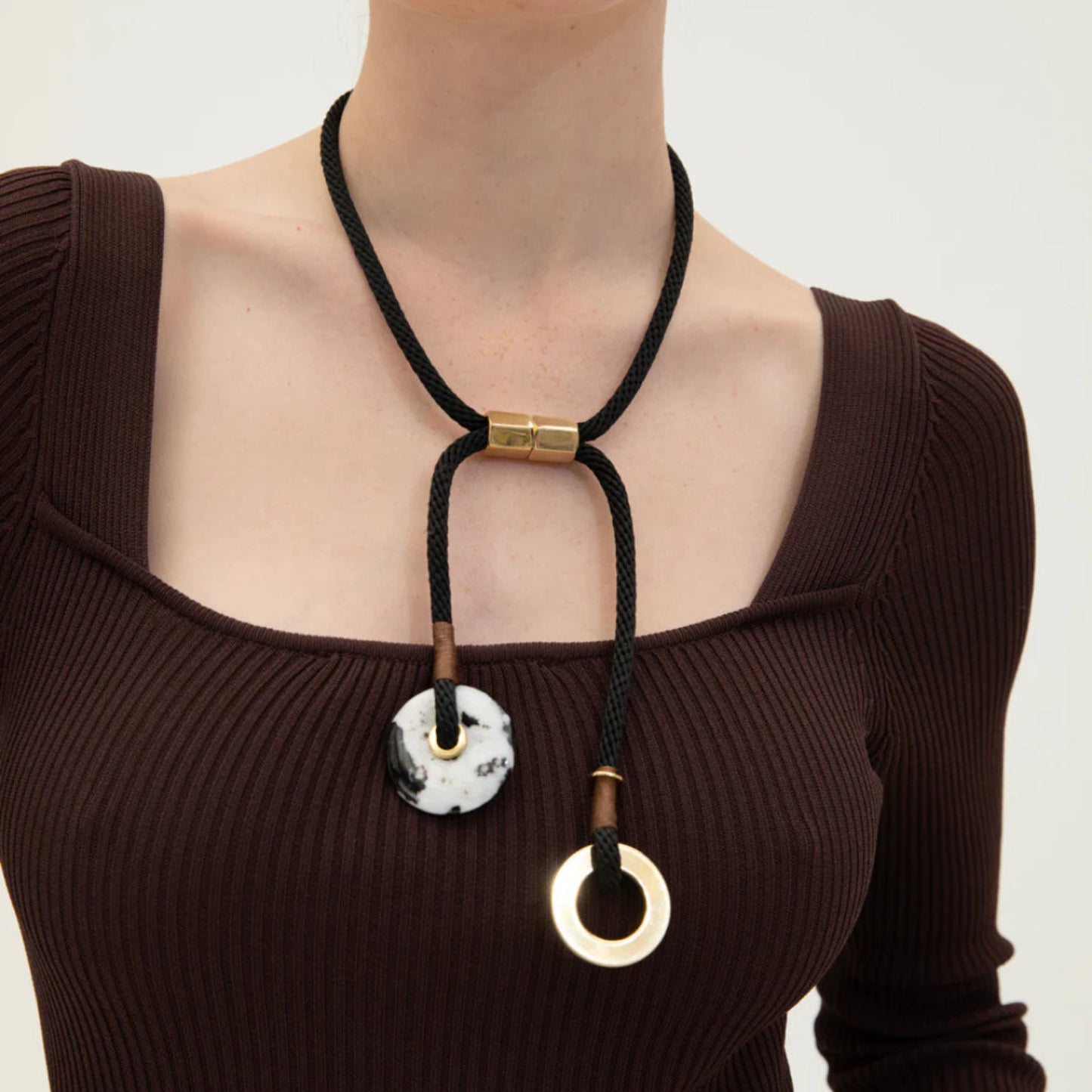 A person wearing a brown top and showcasing a black necklace with a brass magnetic clasp and a zebra jasper stone.