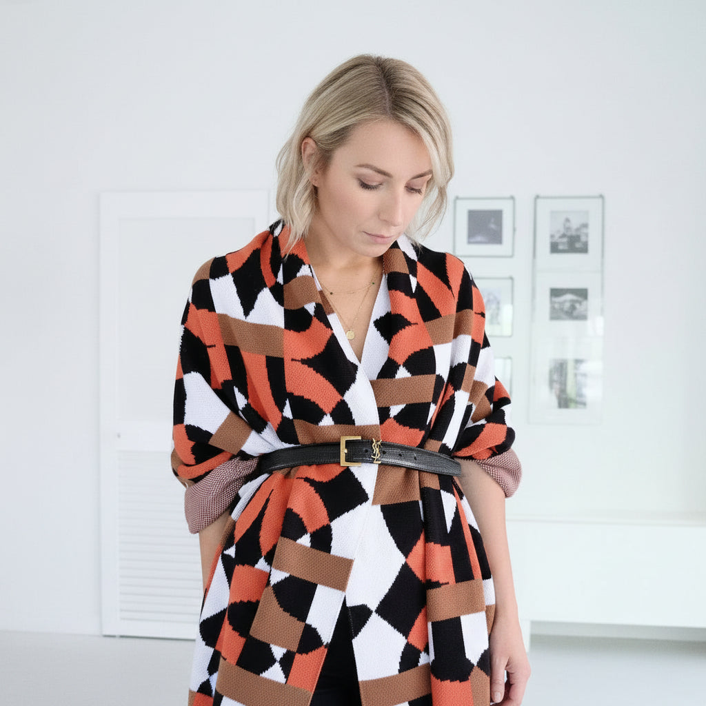 A woman wearing a geometric patterned wrap dress with orange, black, and white stripes, styled with a black belt, standing in a bright white room.