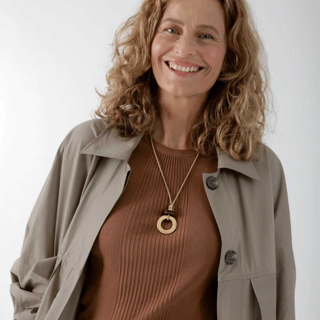 A smiling woman with curly blonde hair wearing a brown ribbed knit top, a beige button-up overshirt, and a gold-tone pendant necklace with a circular ring charm, set against a plain light gray background.