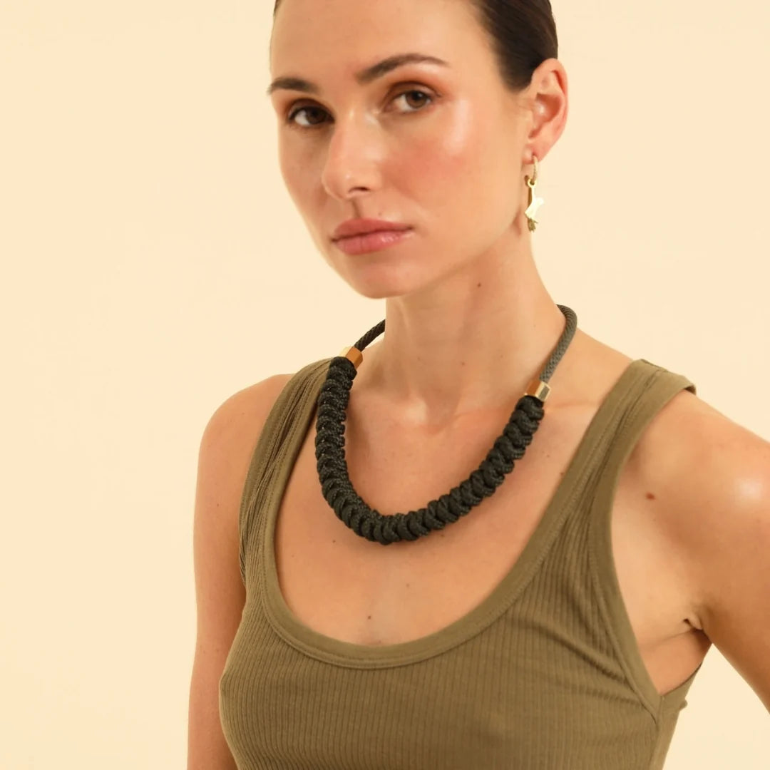 A woman wearing a chunky black braided rope necklace with gold-toned metal accents, paired with an olive green ribbed tank top against a solid light beige background.