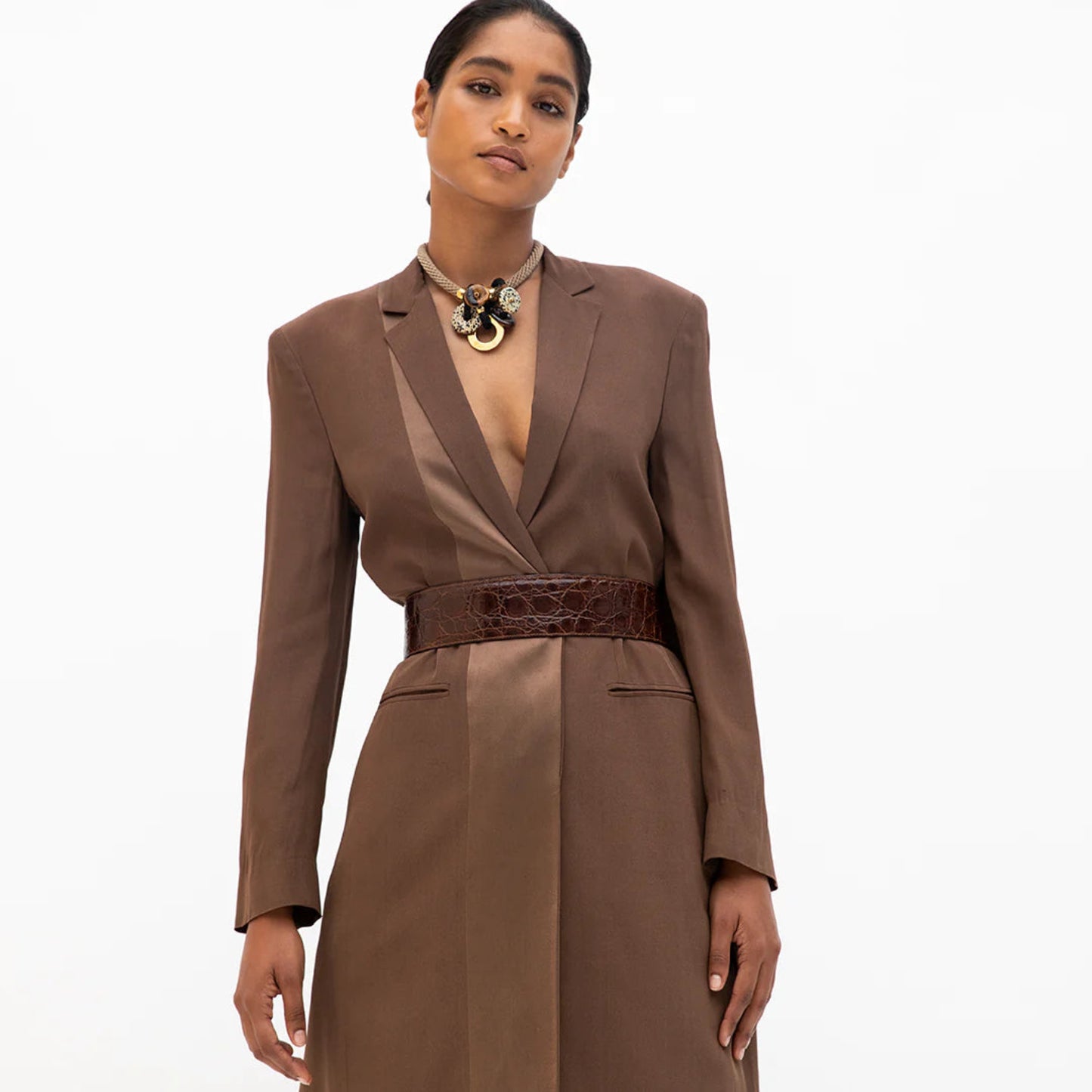 A woman wearing a brown blazer showcasing a beige braided rope necklace embellished with red and dalmatian jasper stones, and brass rings.