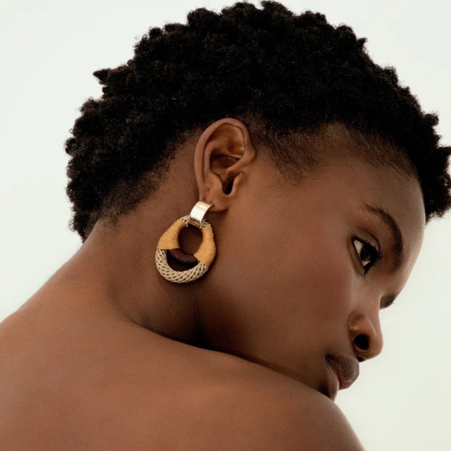 Woman wearing a gold hoop earring with a woven rope in beige, camel and maroon a plain background