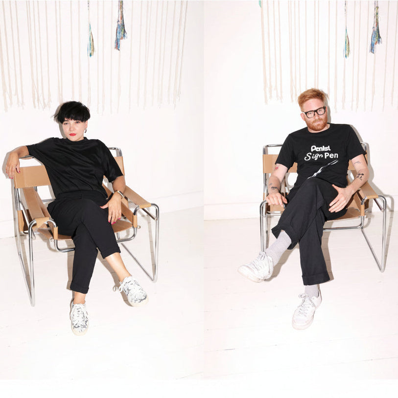Two artist sitting in chairs in a bright white studio setting, both wearing black graphic t-shirts, black pants, and white sneakers. The left model has short black hair and the right model has red hair and glasses.