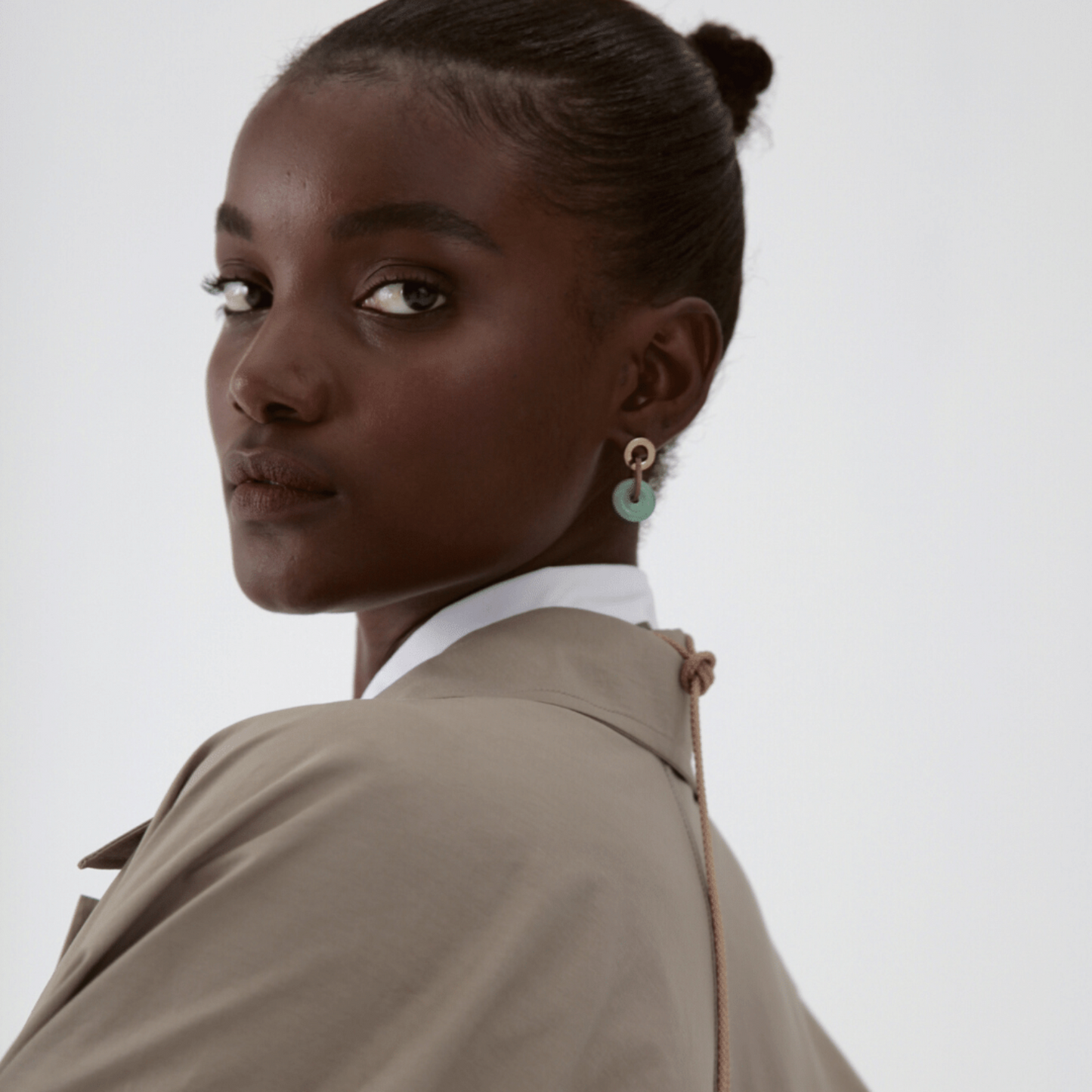 A person wearing a beige coat and green aventurine stone earrings with brass and silver studs.