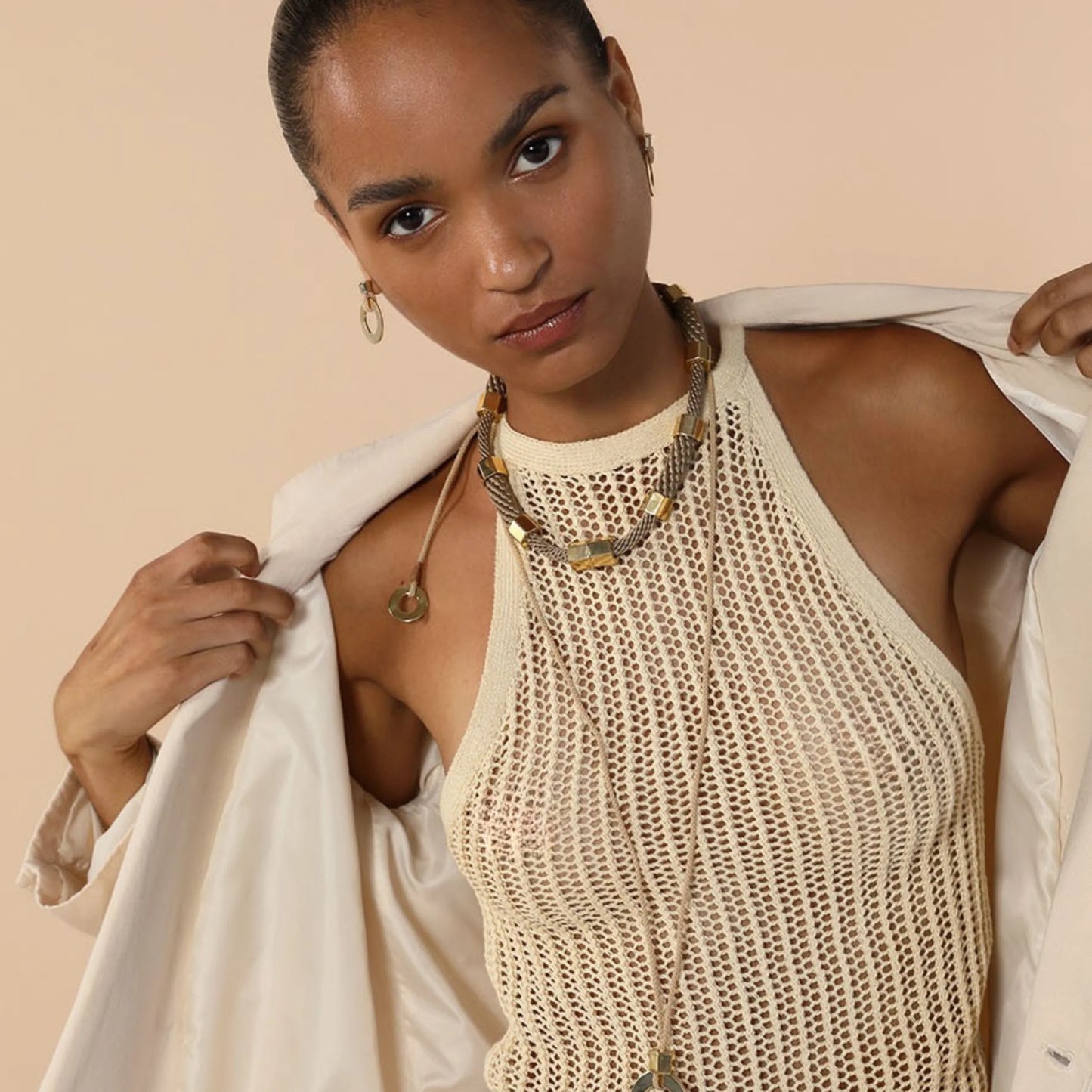 A woman wearing a cream-colored open-knit halter top and holding open a matching cream satin jacket against a solid beige background.