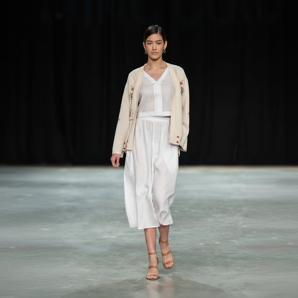 Model walking on a runway wearing a light-colored outfit with a white top and wide-leg pants.