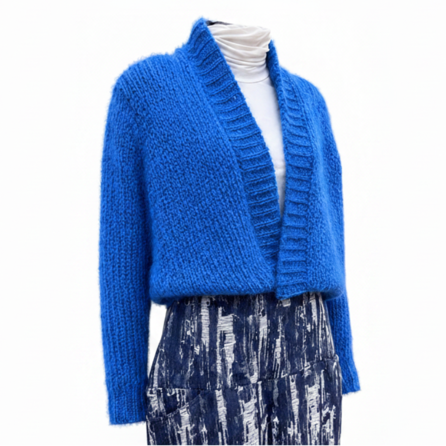 A bright blue chunky knit cardigan sweater displayed on a mannequin torso, layered over a white turtleneck and a blue-and-white patterned skirt, against a plain white background.