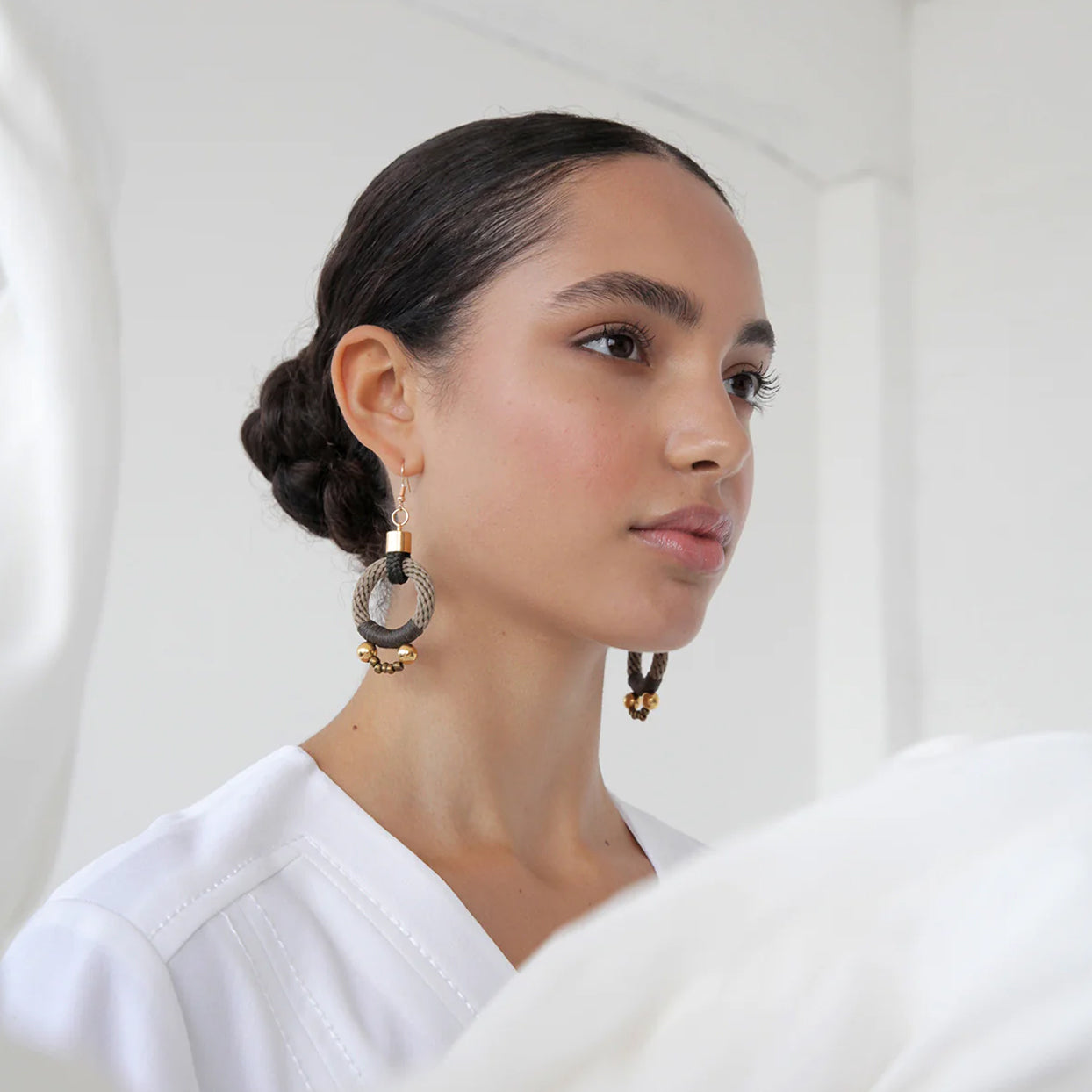 A gorgeous woman stares at herself in the mirror. She is wearing the PICHULIK Alpha Earrings with a sleek bun. The olive and beige tone looks striking against her warm skin tone.