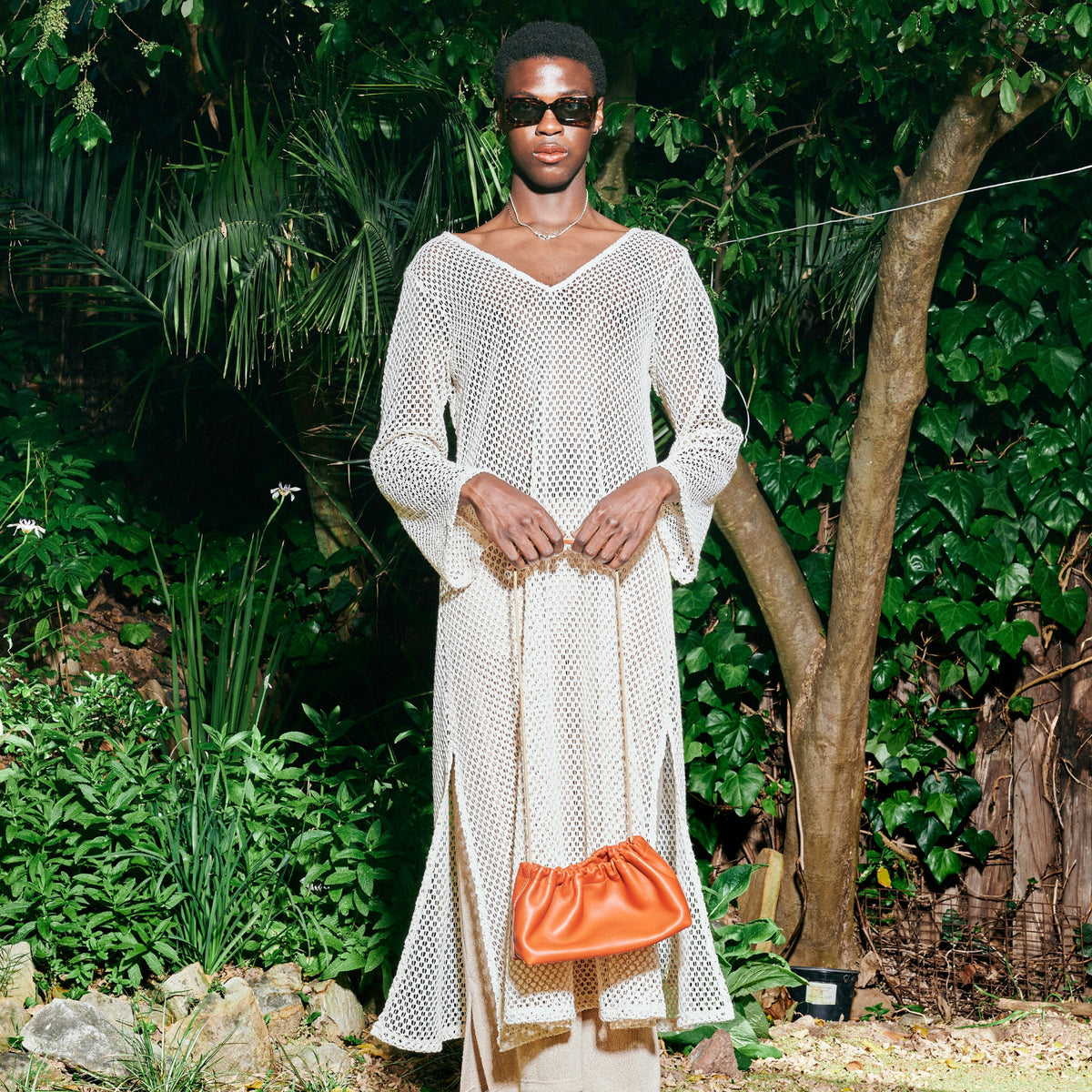 Person wearing a white knitted dress and sunglasses, holding a brown handbag in a garden setting.