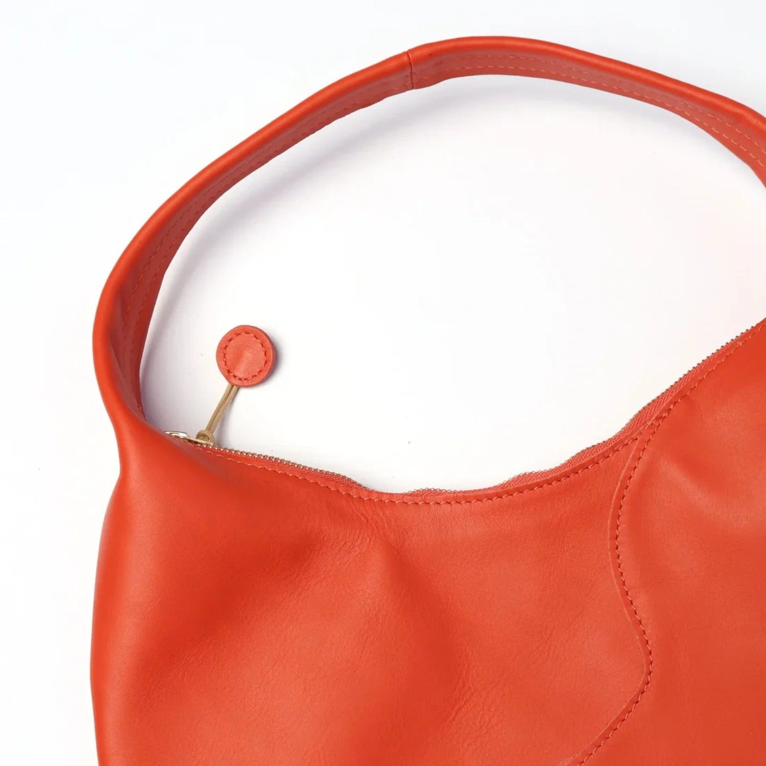 Close-up of a bright orange-red leather shoulder bag with a curved top handle, zipper closure, and a small matching leather pull tab on a white background.