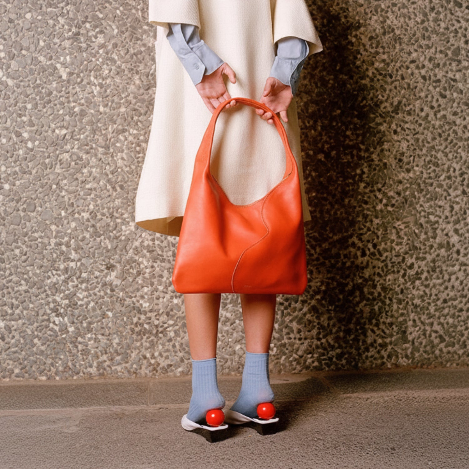 A person wearing a cream dress and light blue shirt holds a large orange leather hobo bag. The person is also wearing light blue socks and black wedge sandals with red balls on the soles, standing against a textured stone wall.