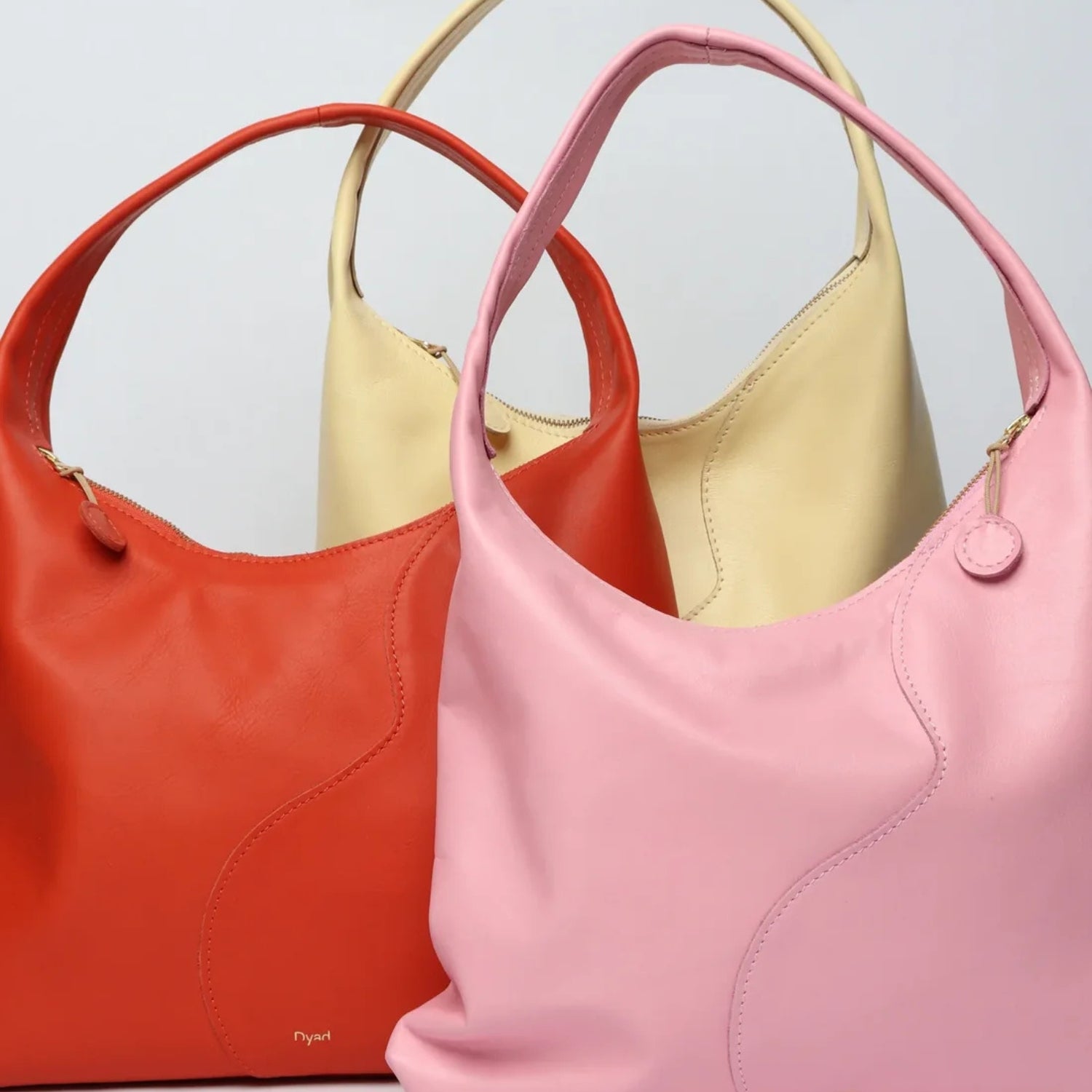 Three leather shoulder bags in coral red, pale yellow, and light pink, arranged overlapping against a light gray background.