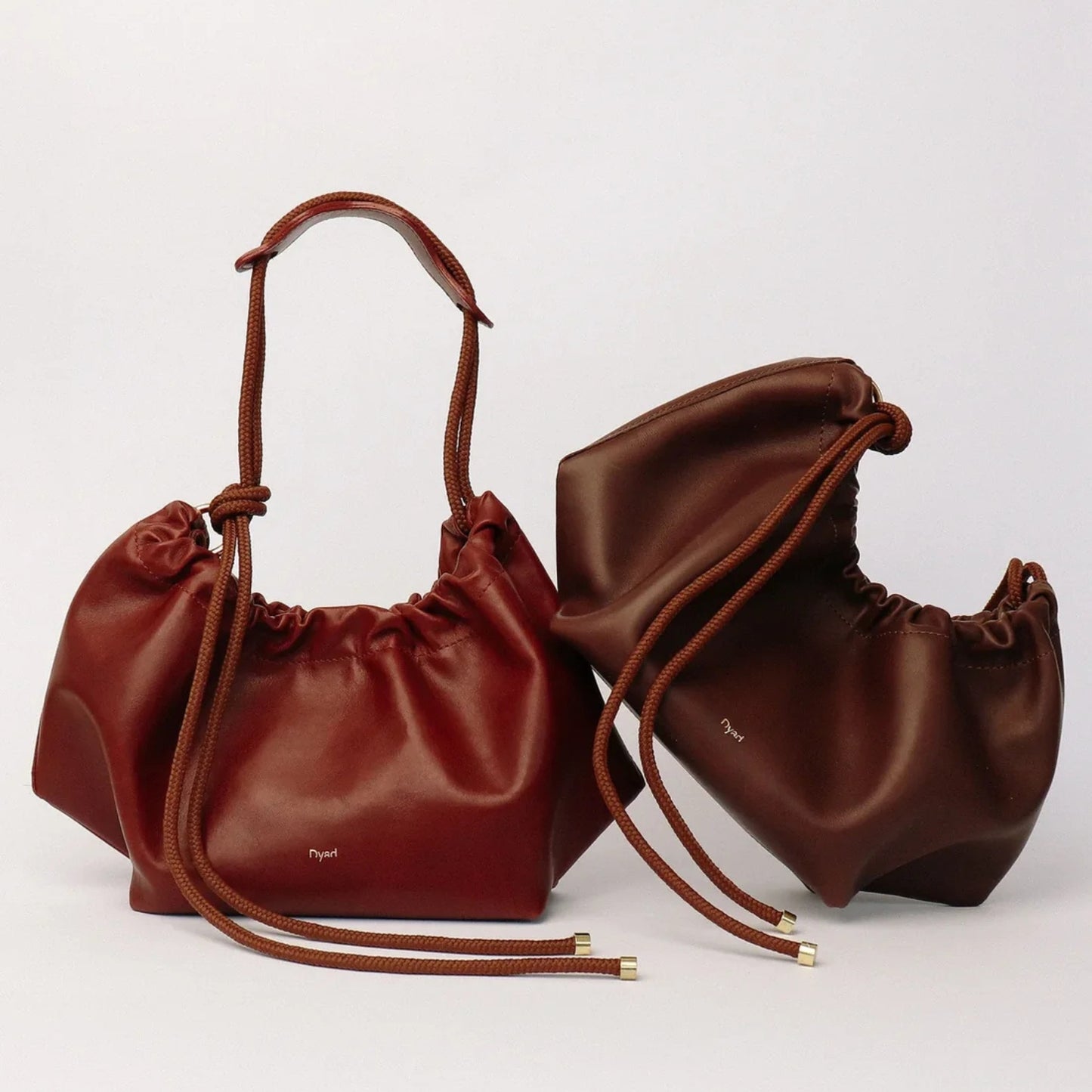 Two small drawstring pouch bags in rich reddish-brown leather, one standing upright and the other lying on its side, both with matching rope handles and gold-tone tip details, photographed on a clean light gray background.