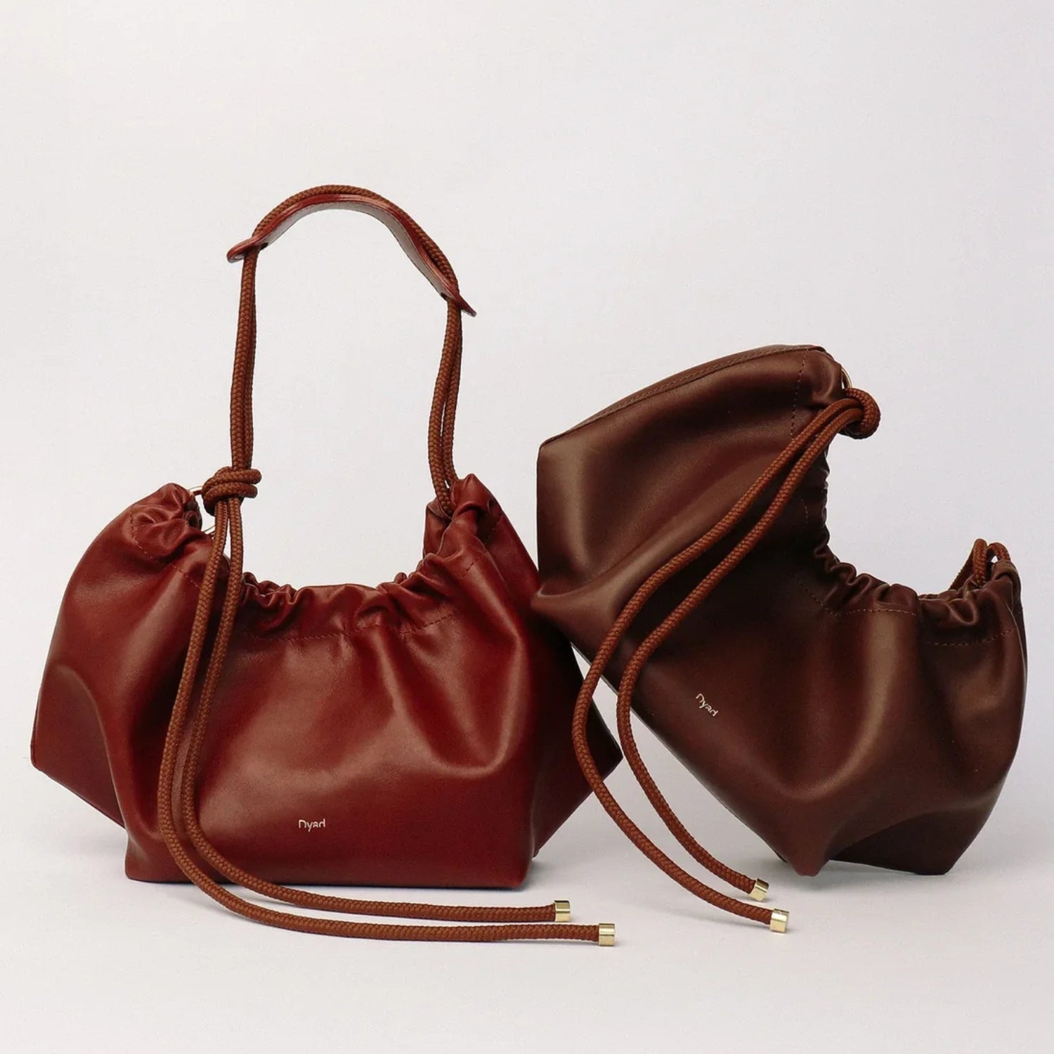 Two small drawstring pouch bags in rich reddish-brown leather, one standing upright and the other lying on its side, both with matching rope handles and gold-tone tip details, photographed on a clean light gray background.