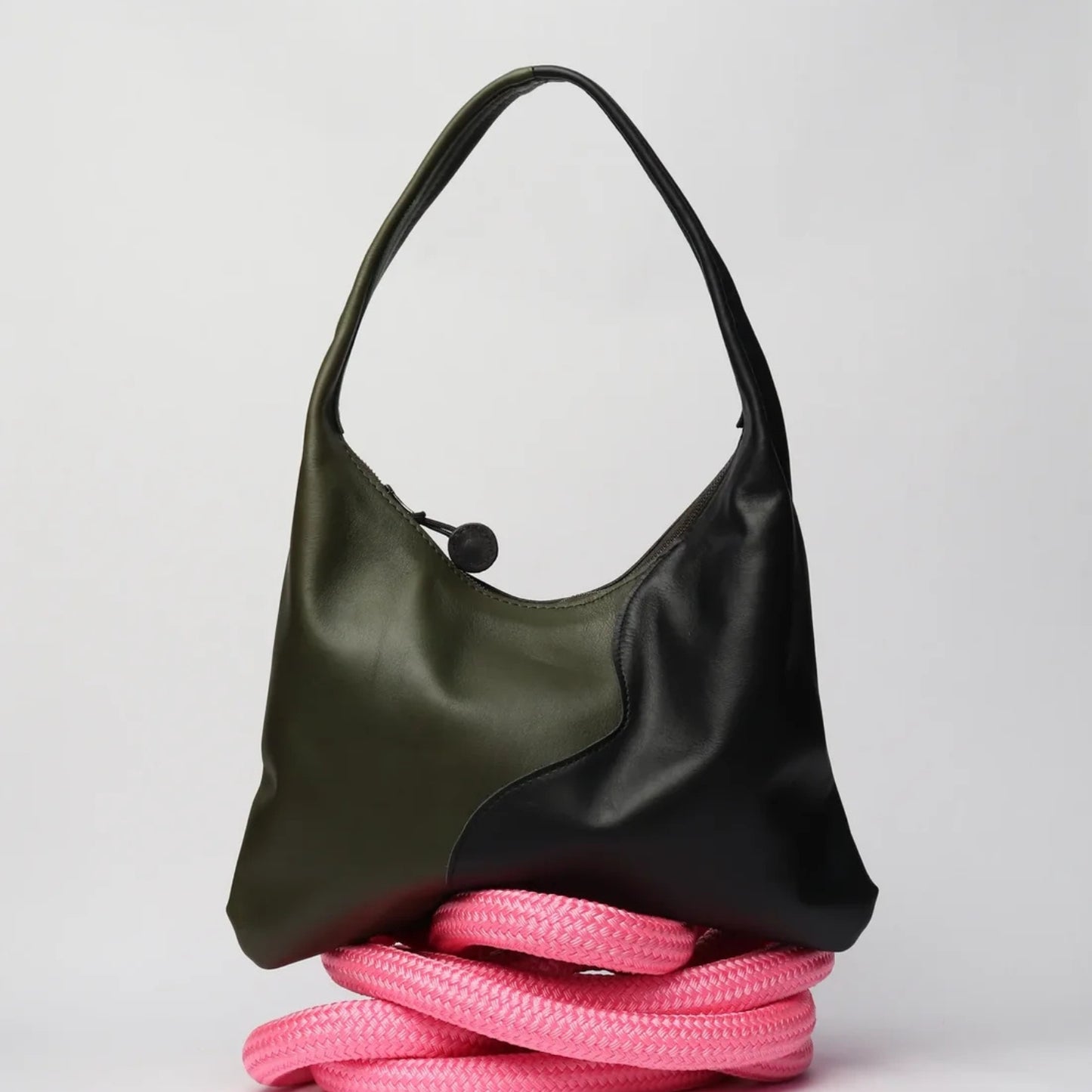 A color-blocked hobo shoulder bag with an olive green leather half and a black leather half, resting on a coiled pink rope against a light gray background.