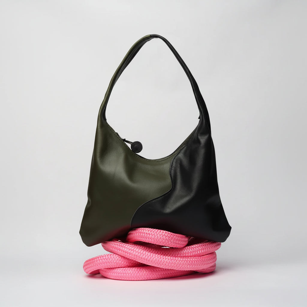 A color-blocked hobo shoulder bag with an olive green leather half and a black leather half, resting on a coiled pink rope against a light gray background.