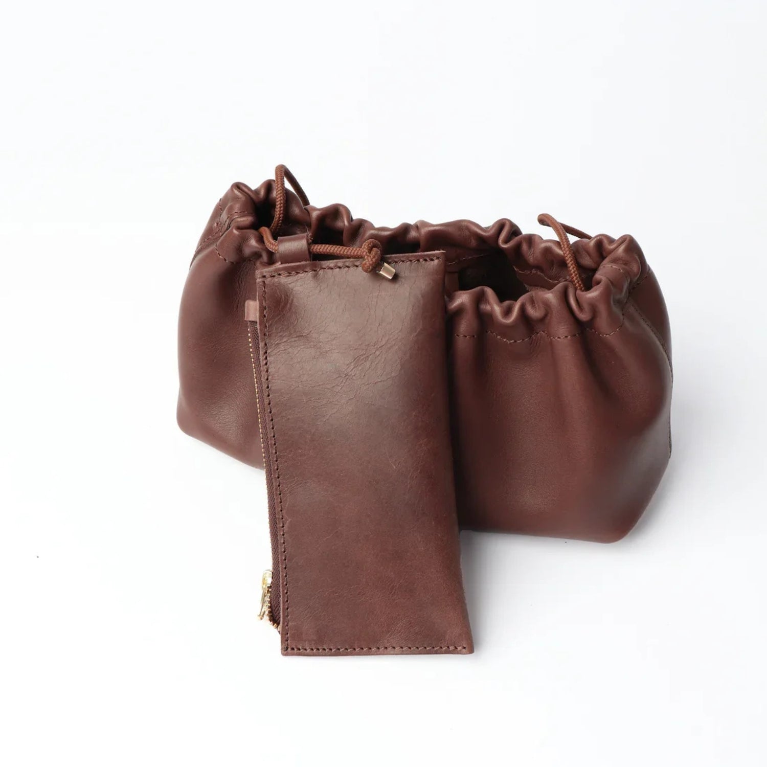 A brown leather accessory set featuring a drawstring pouch, a matching zippered pouch, and a small leather card holder with gold-tone hardware, displayed on a clean white background.