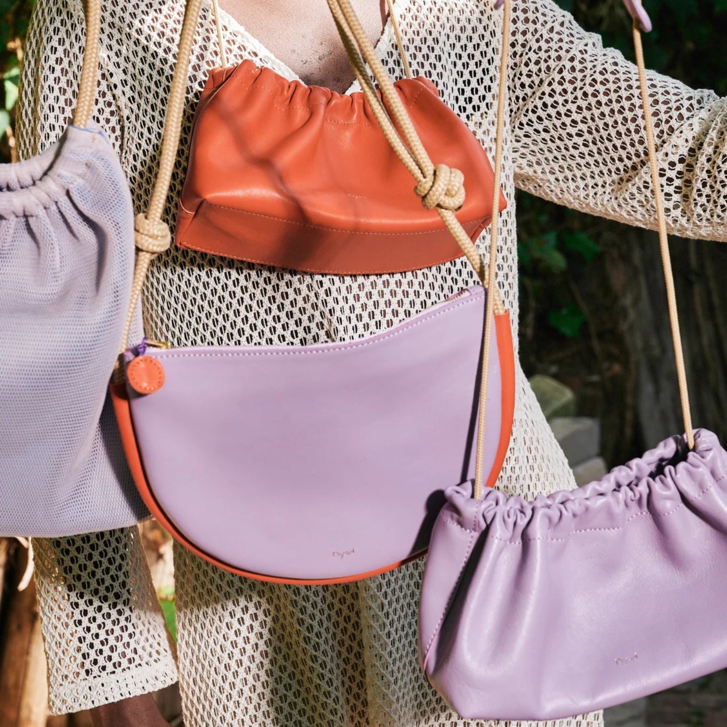 Three leather and woven crossbody bags in lavender, Orange-red, and light purple with orange trim, shown hanging from a person's shoulders outdoors.