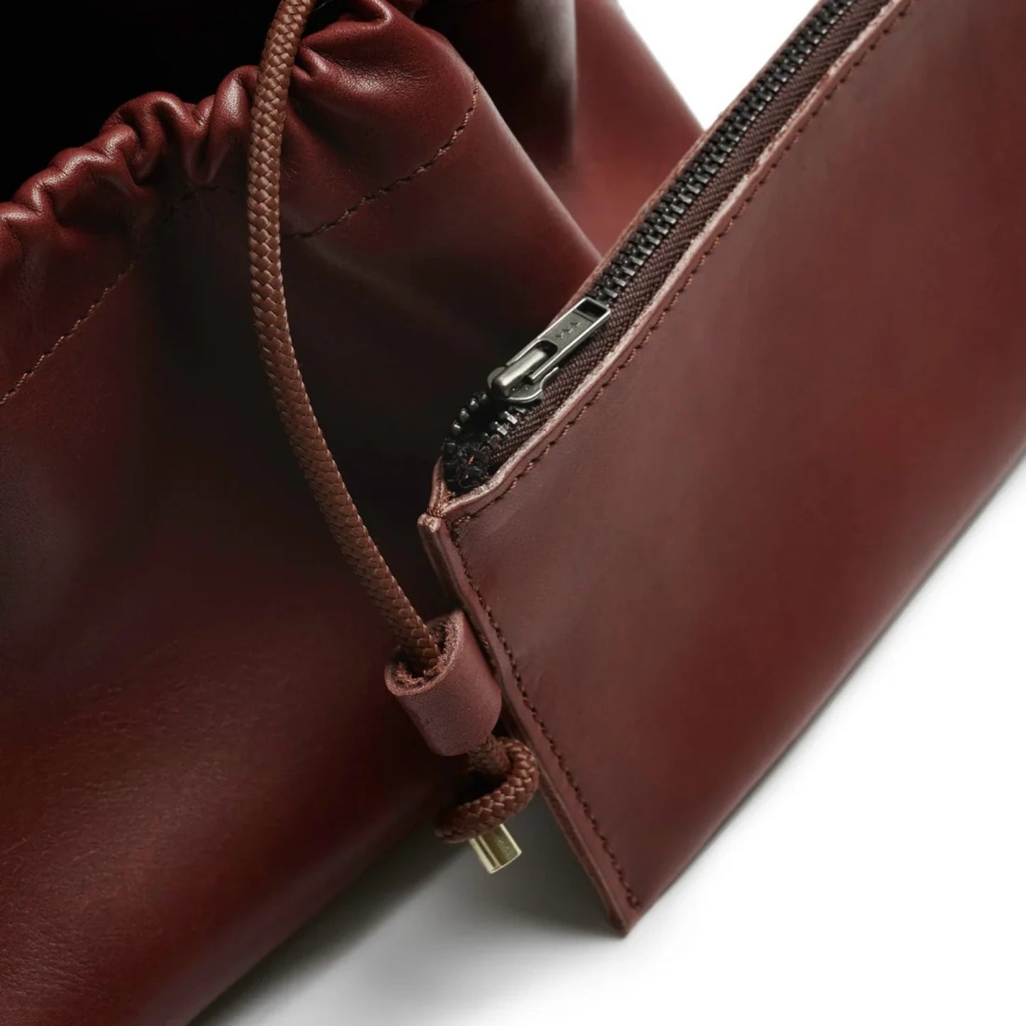 Close-up of a dark reddish-brown leather bag showing a drawstring closure, a zippered pocket, and a bronze zipper pull on a white background.