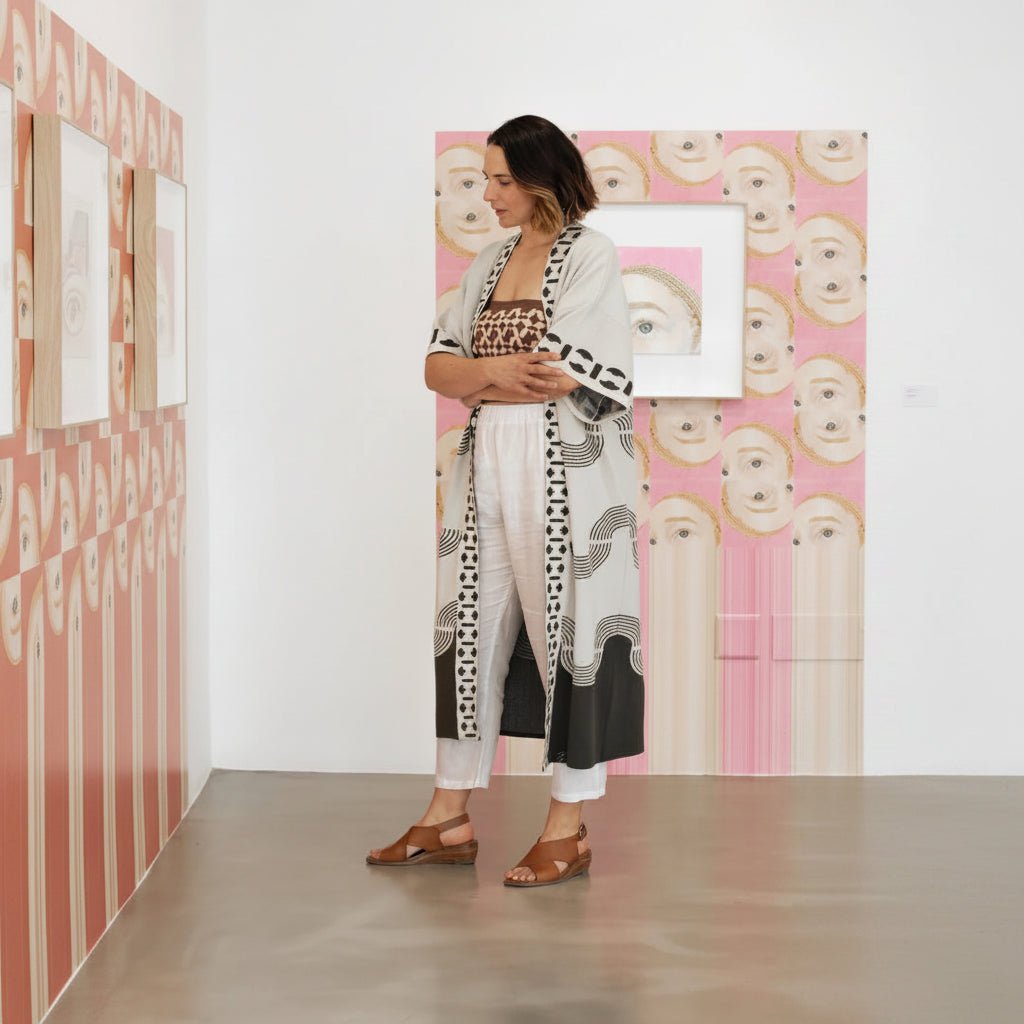 A woman stands in a gallery wearing a long open-front cardigan with black, white, and gray abstract geometric patterns over a leopard-print top and white pants. The background features pink and white art pieces.