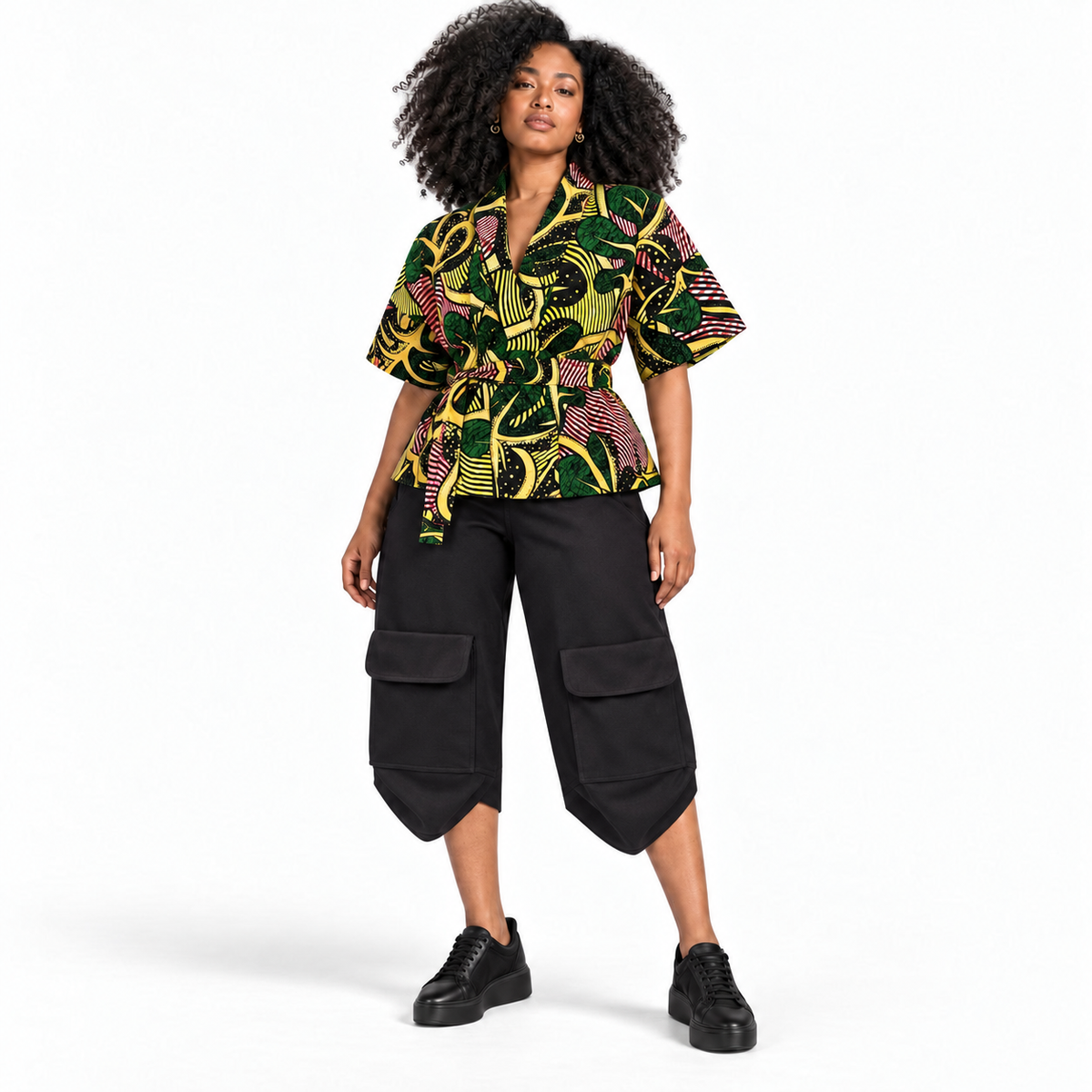 A model wearing a short-sleeve top with a vibrant green, yellow, and pink abstract pattern, paired with black cargo cropped pants and black platform sneakers, set against a plain white studio background.