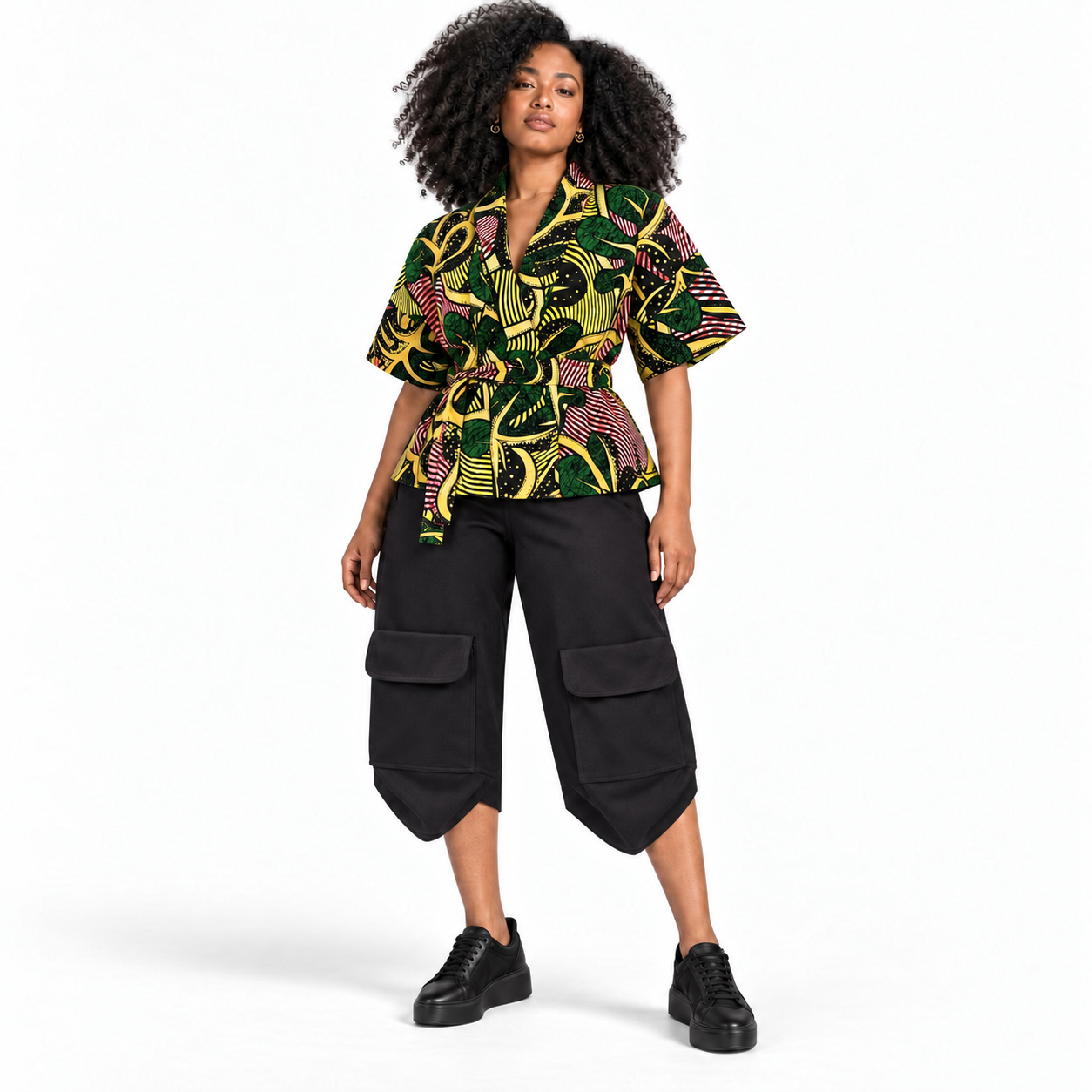 A model wearing a short-sleeve top with a vibrant green, yellow, and pink abstract pattern, paired with black cargo cropped pants and black platform sneakers, set against a plain white studio background.