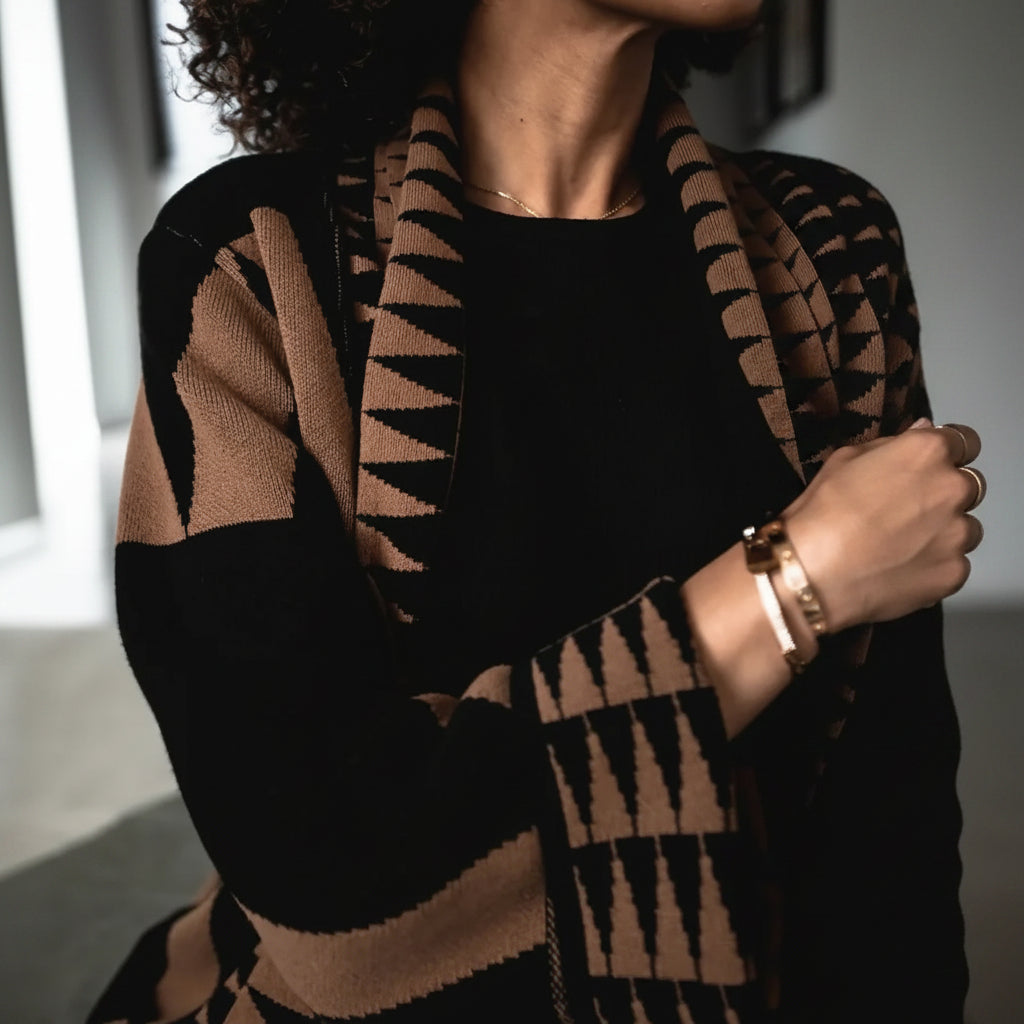 A close-up of a person wearing a black knit cardigan with tan geometric patterns and a matching knit scarf, styled over a black top.