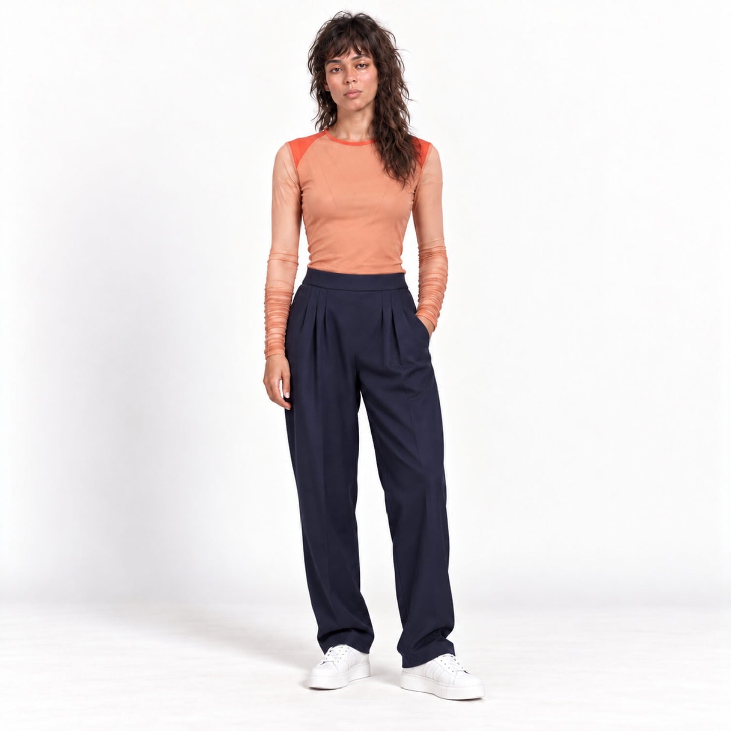 A model stands against a white studio background wearing a peach long-sleeve top with orange shoulder panels and dark navy pleated trousers, paired with white sneakers.