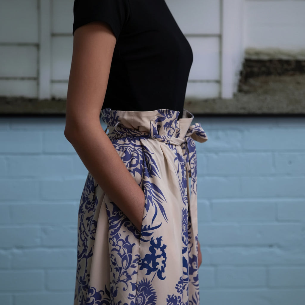 A close-up of a person wearing a beige paperbag-style shorts with a blue floral damask pattern, paired with a black top, set against a light blue brick wall.