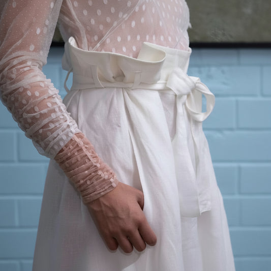 A close-up of a white linen dress with a matching fabric belt and bow, worn by a model whose hand and arm in a sheer pink lace sleeve are visible.