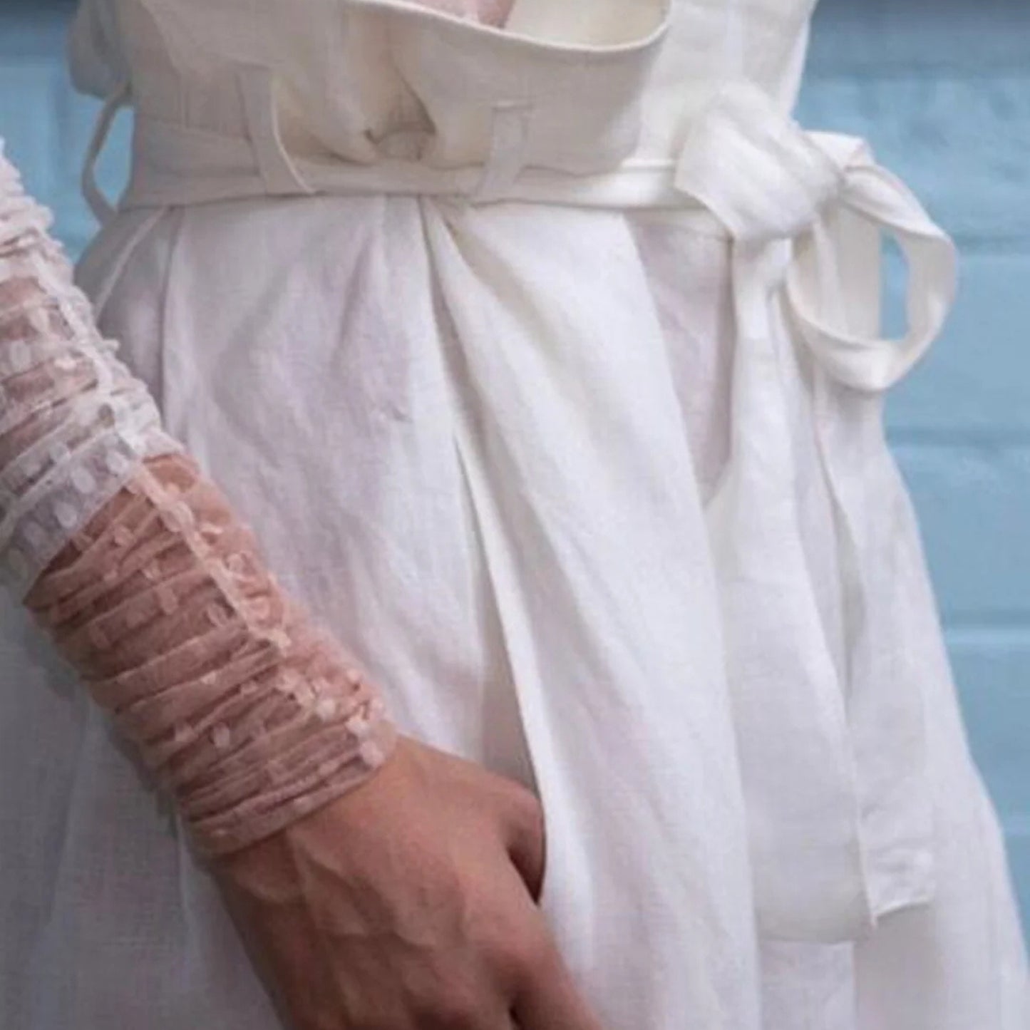 A close-up of a white linen dress with a matching fabric belt and bow, worn by a model whose hand and arm in a sheer pink lace sleeve are visible.
