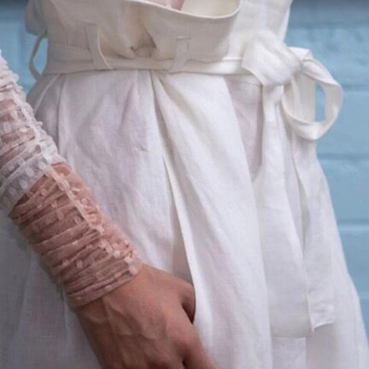 A close-up of a white linen dress with a matching fabric belt and bow, worn by a model whose hand and arm in a sheer pink lace sleeve are visible.
