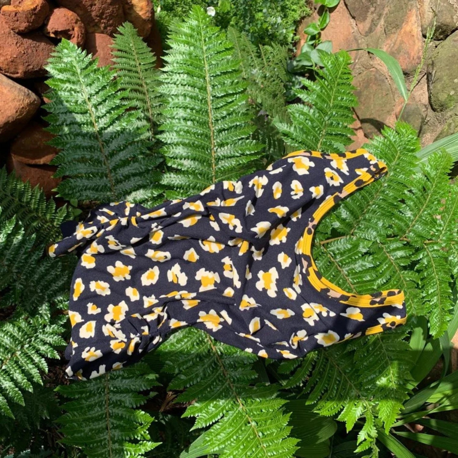 A black sleeveless top with a yellow and white floral pattern, laid flat on a bed of green fern leaves.