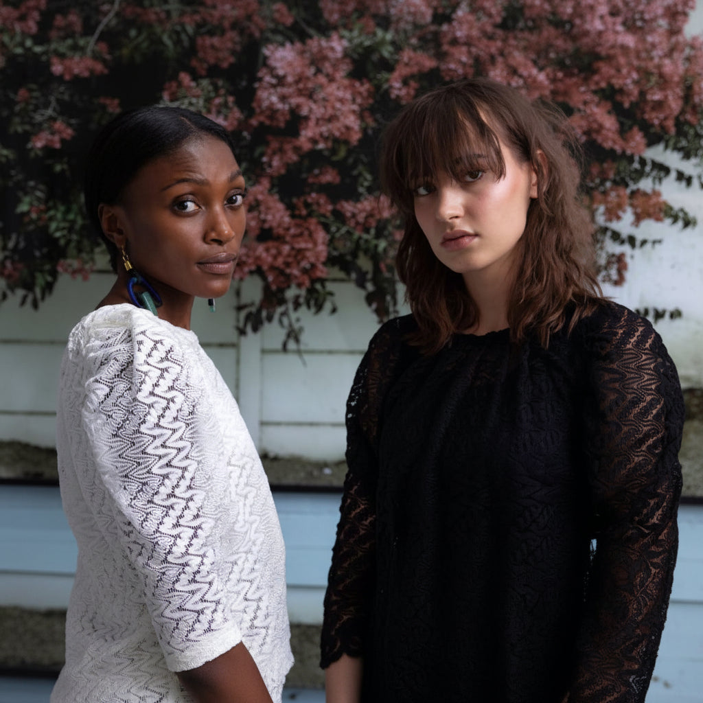 Two women posing outdoors in front of a white wall and pink flowers. The woman on the left wears a white textured top and blue earrings, while the woman on the right wears a black lace top.