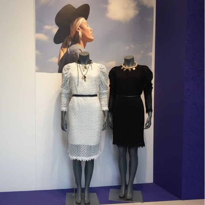 Two long-sleeve lace dresses displayed on mannequins indoors. The left dress is white with a black waistband and layered necklaces, and the right dress is black with a gold necklace.
