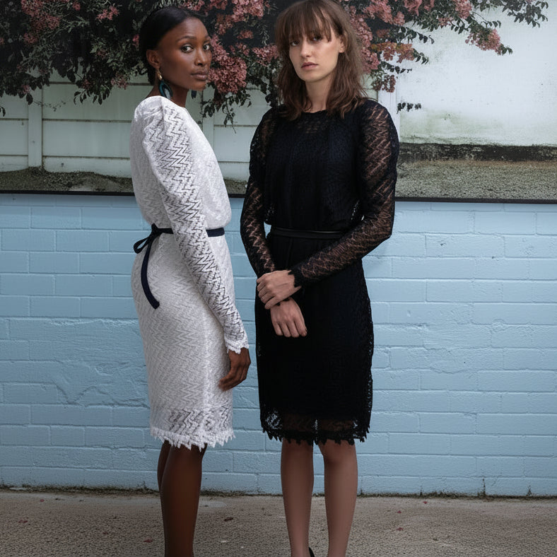 Two women modeling long-sleeve lace dresses, one in white and one in black, standing against a light blue brick wall with foliage in the background.