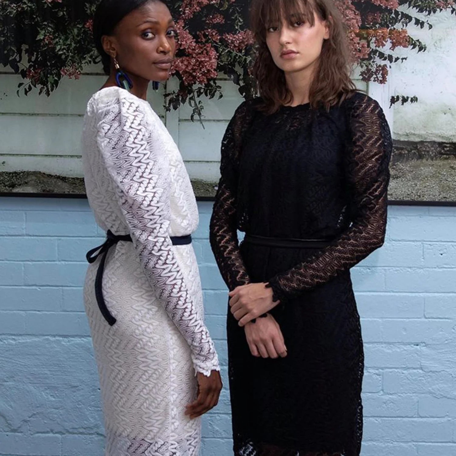 Two women modeling long-sleeve lace dresses, one in white and one in black, standing against a light blue brick wall with foliage in the background.