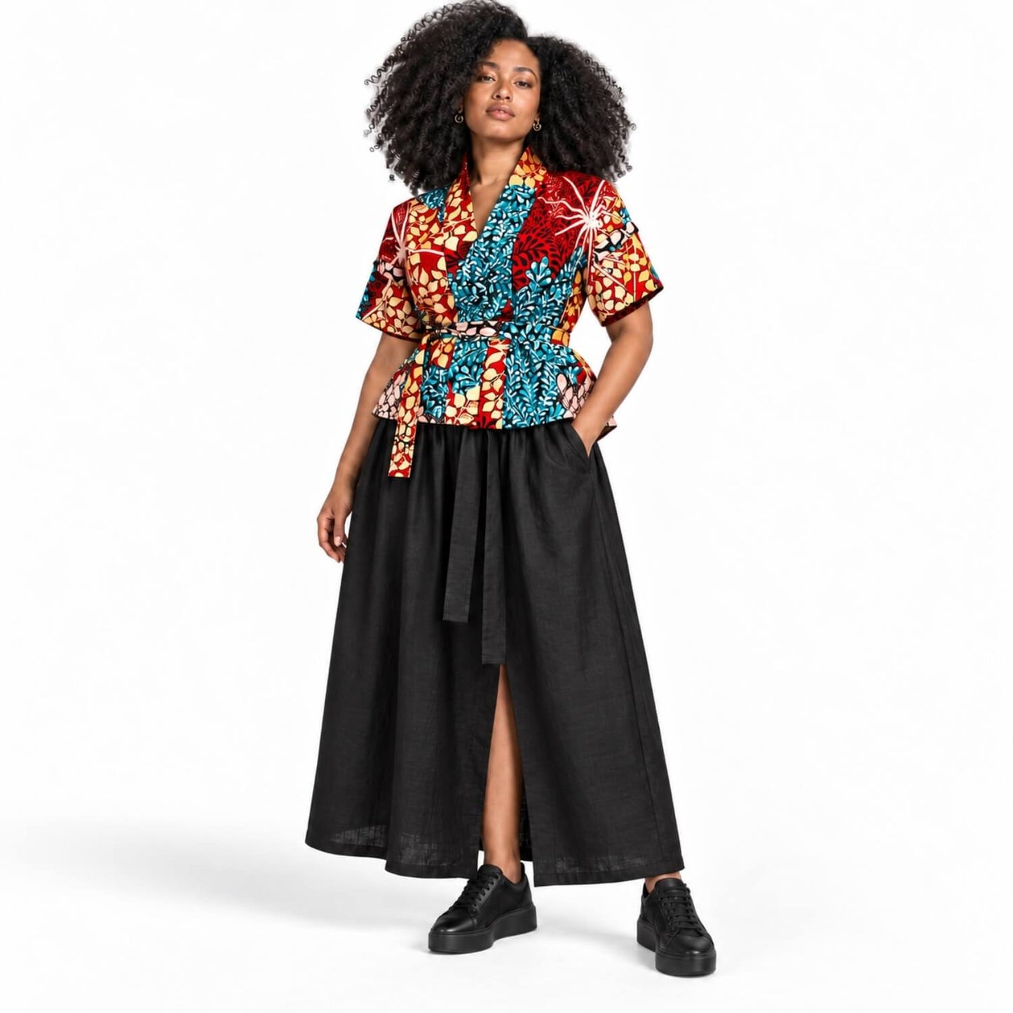A model with curly black hair is wearing a short-sleeve wrap-style top with a vibrant multicolor African wax print (red, blue, orange, yellow) paired with a long black skirt and black platform sneakers, shot against a solid white studio background.