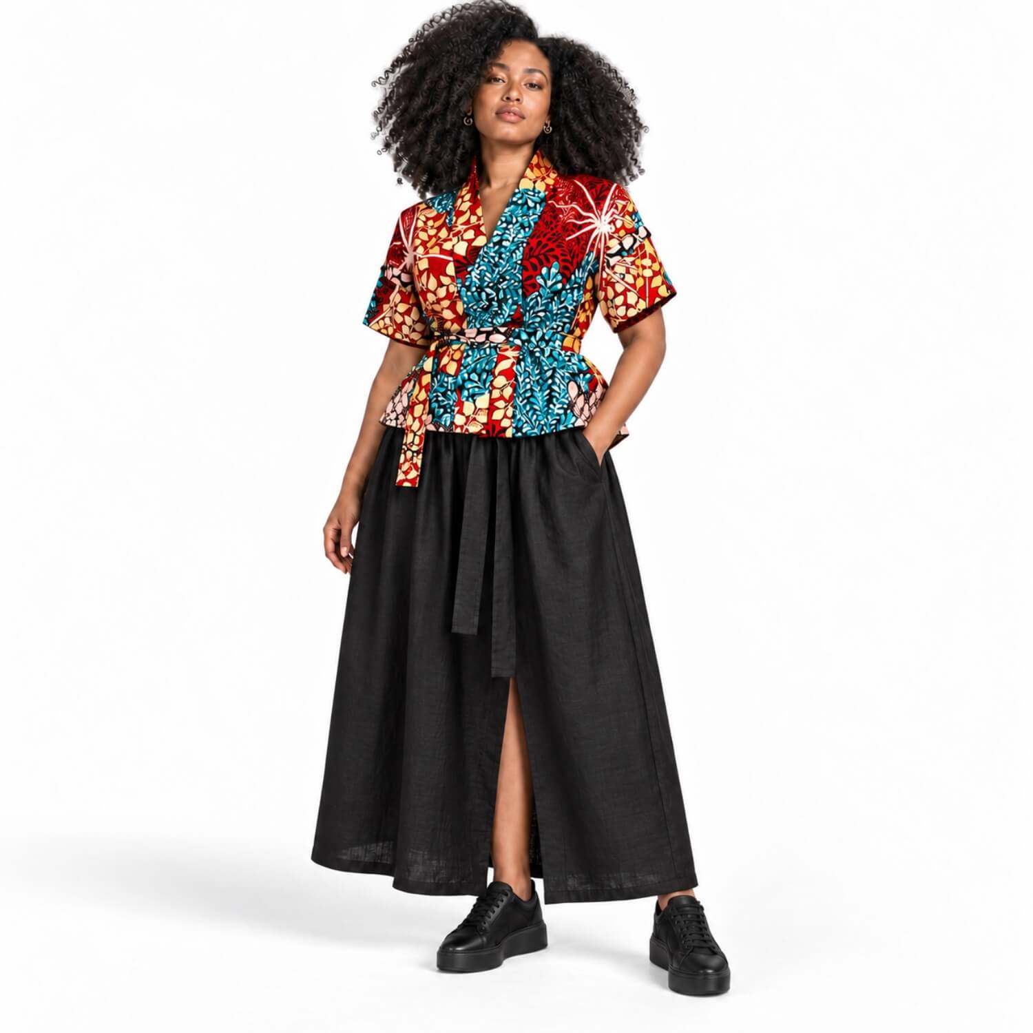 A model with curly black hair is wearing a short-sleeve wrap-style top with a vibrant multicolor African wax print (red, blue, orange, yellow) paired with a long black skirt and black platform sneakers, shot against a solid white studio background.