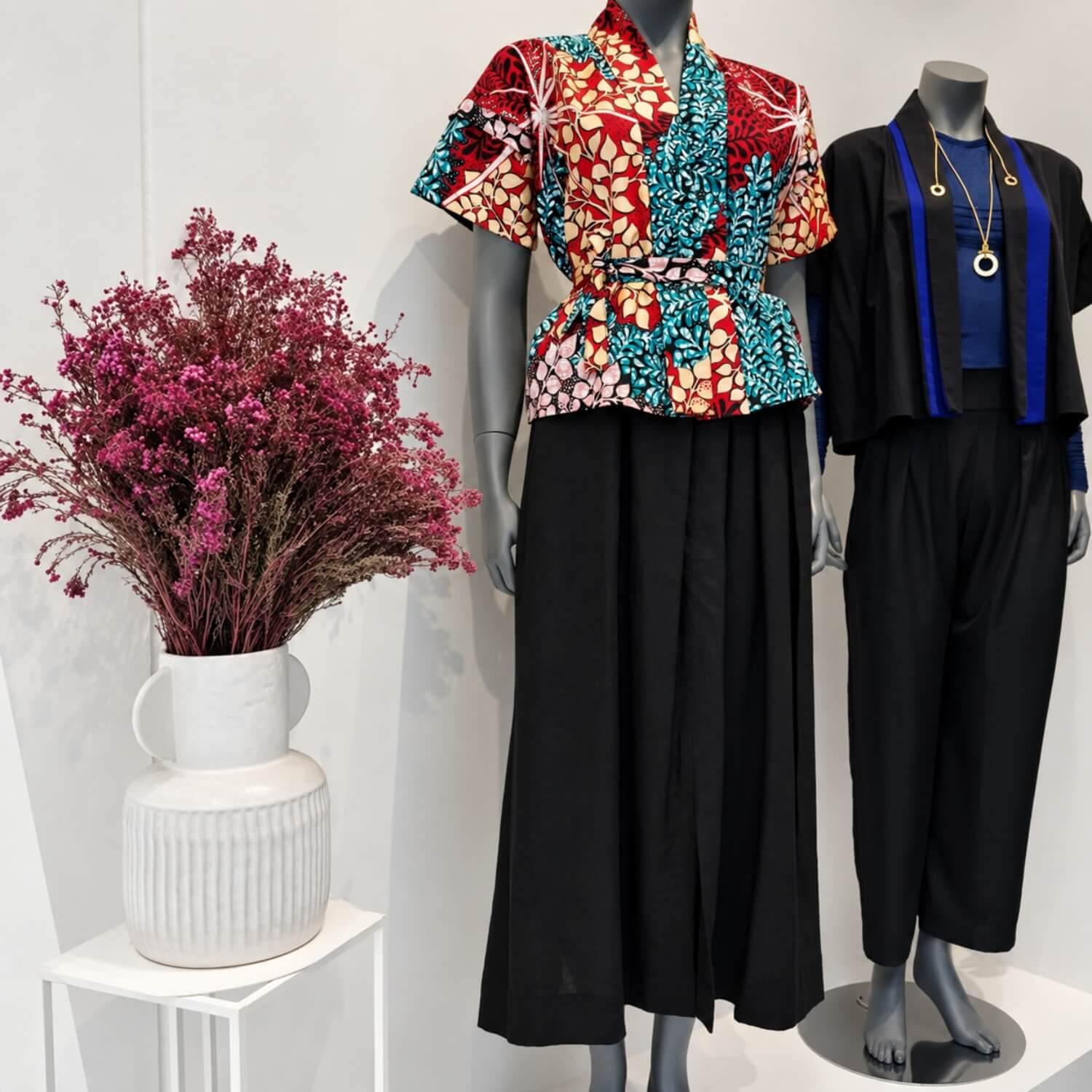 Two mannequins display fashion outfits against a white wall. The left mannequin wears a short-sleeve peplum top with a bold red, teal, and white floral pattern paired with a black skirt.