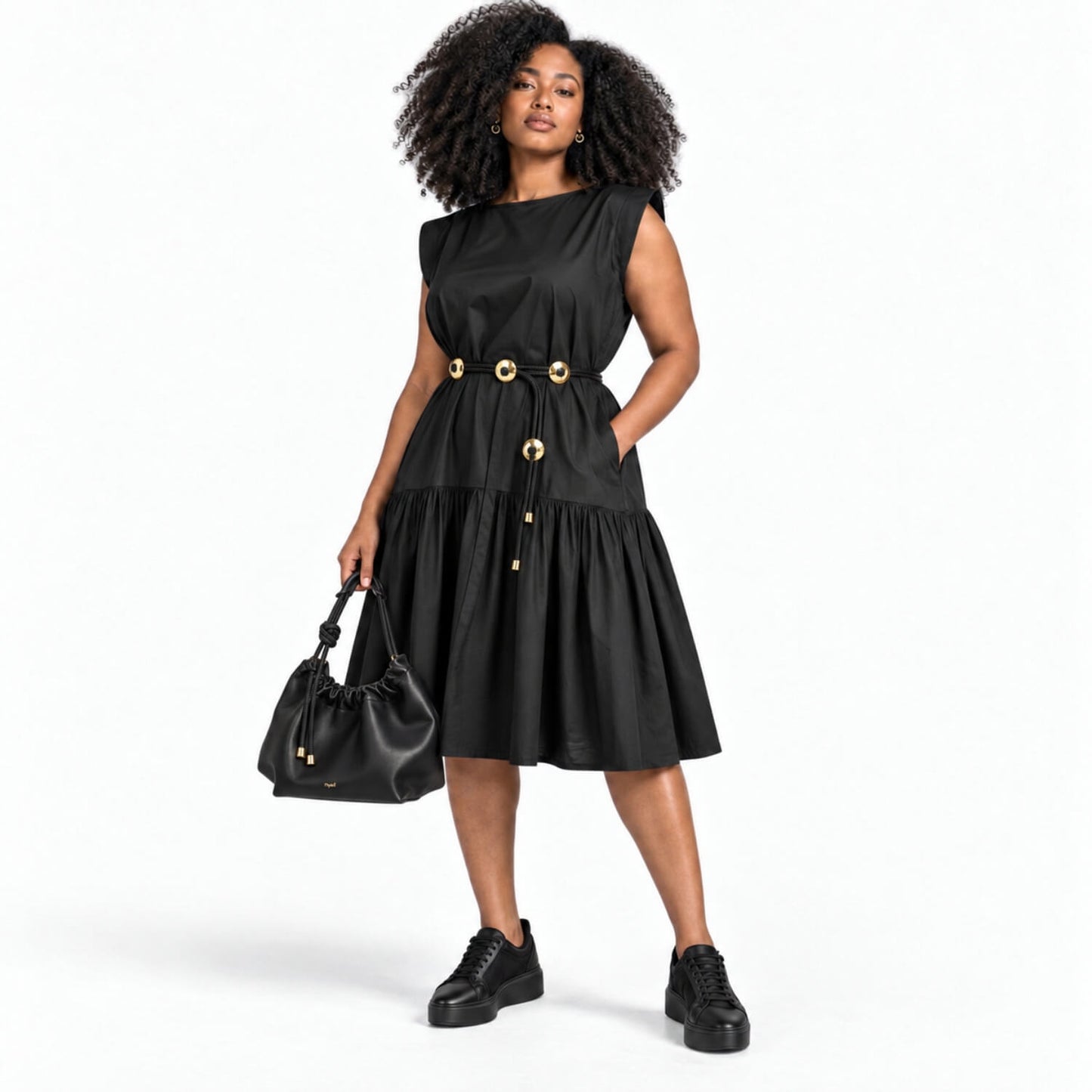 A woman modeling a black sleeveless midi dress with gold-tone decorative buttons and a drawstring waist, paired with black platform sneakers and a black handbag, against a plain white studio background.