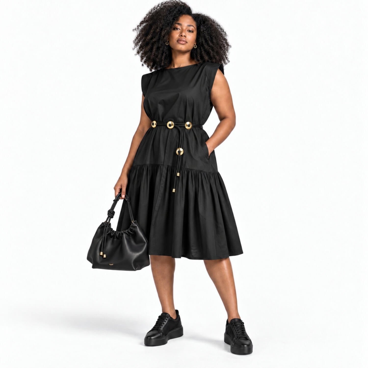 A woman modeling a black sleeveless midi dress with gold-tone decorative buttons and a drawstring waist, paired with black platform sneakers and a black handbag, against a plain white studio background.