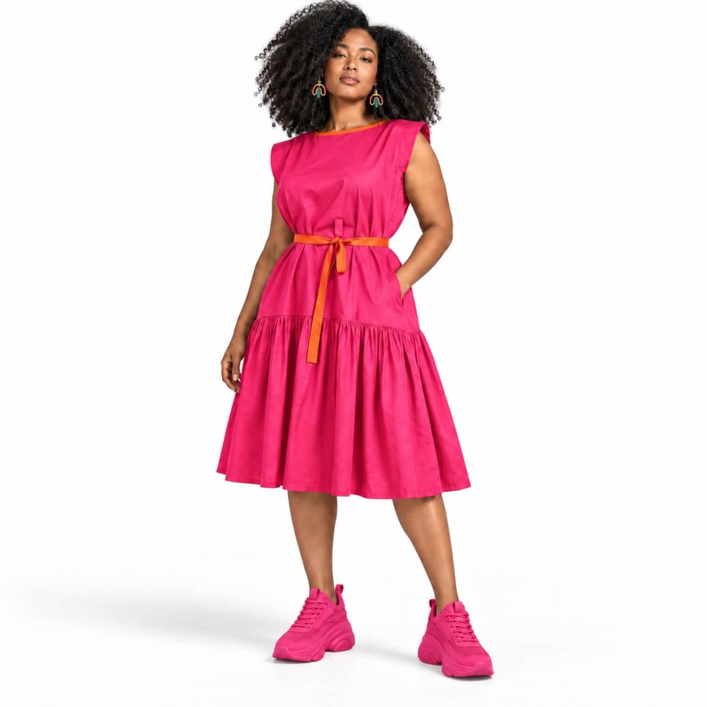 A woman modeling a bright pink sleeveless tiered midi dress with a contrasting orange waist tie belt, paired with matching pink sneakers, against a plain white studio background.