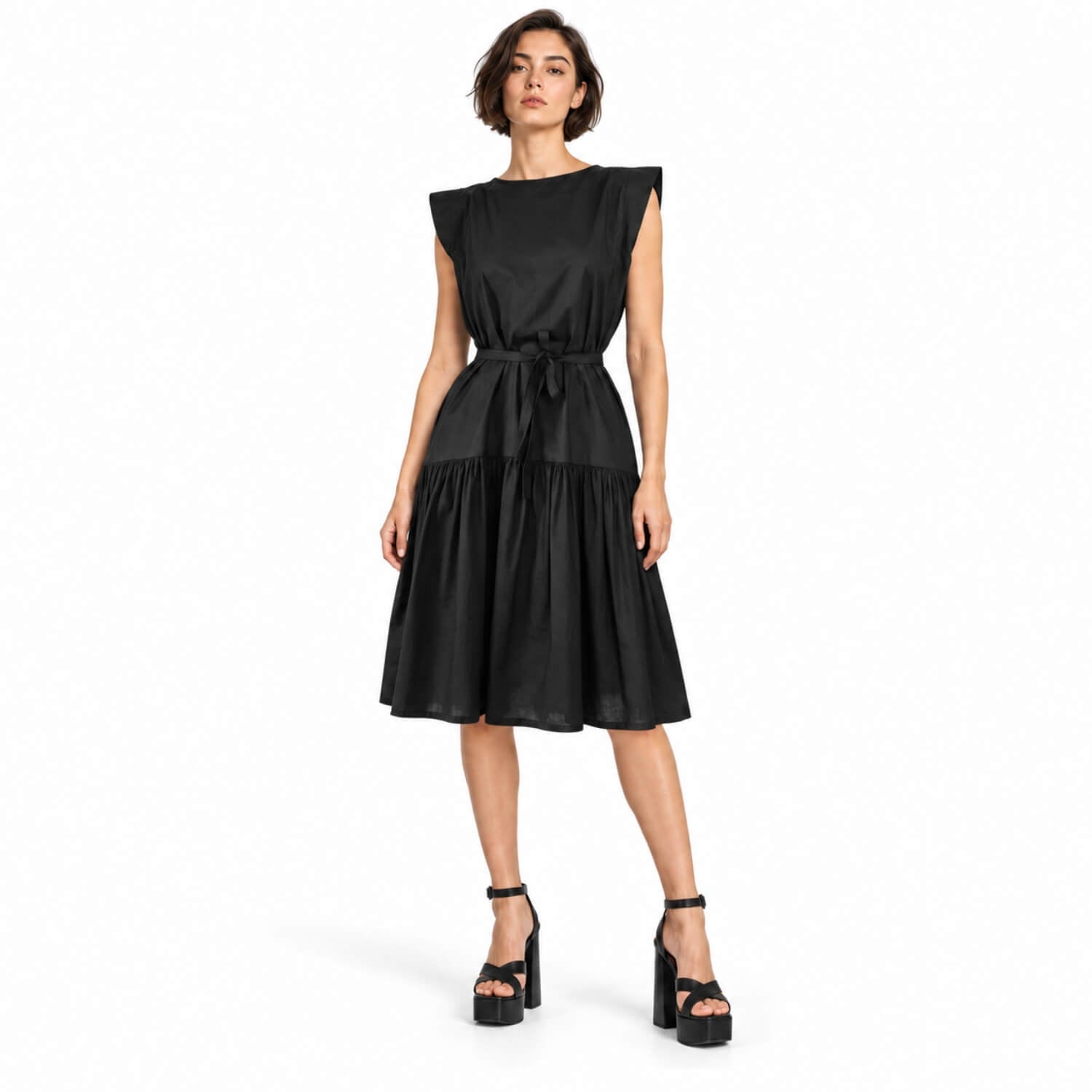A woman modeling a black sleeveless midi dress with a tie waist and tiered skirt, paired with black platform sandals, against a plain white background.