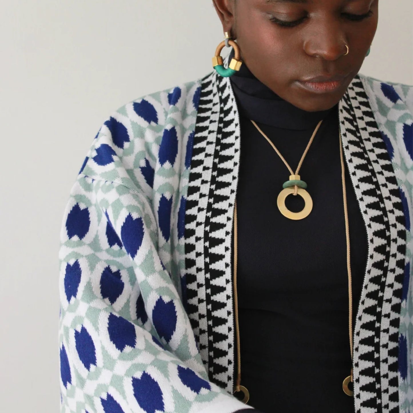 A close-up of a person wearing a navy turtleneck, a patterned knit cardigan with blue, white, and black motifs, and gold jewelry including a large gold pendant necklace and hoop earrings.