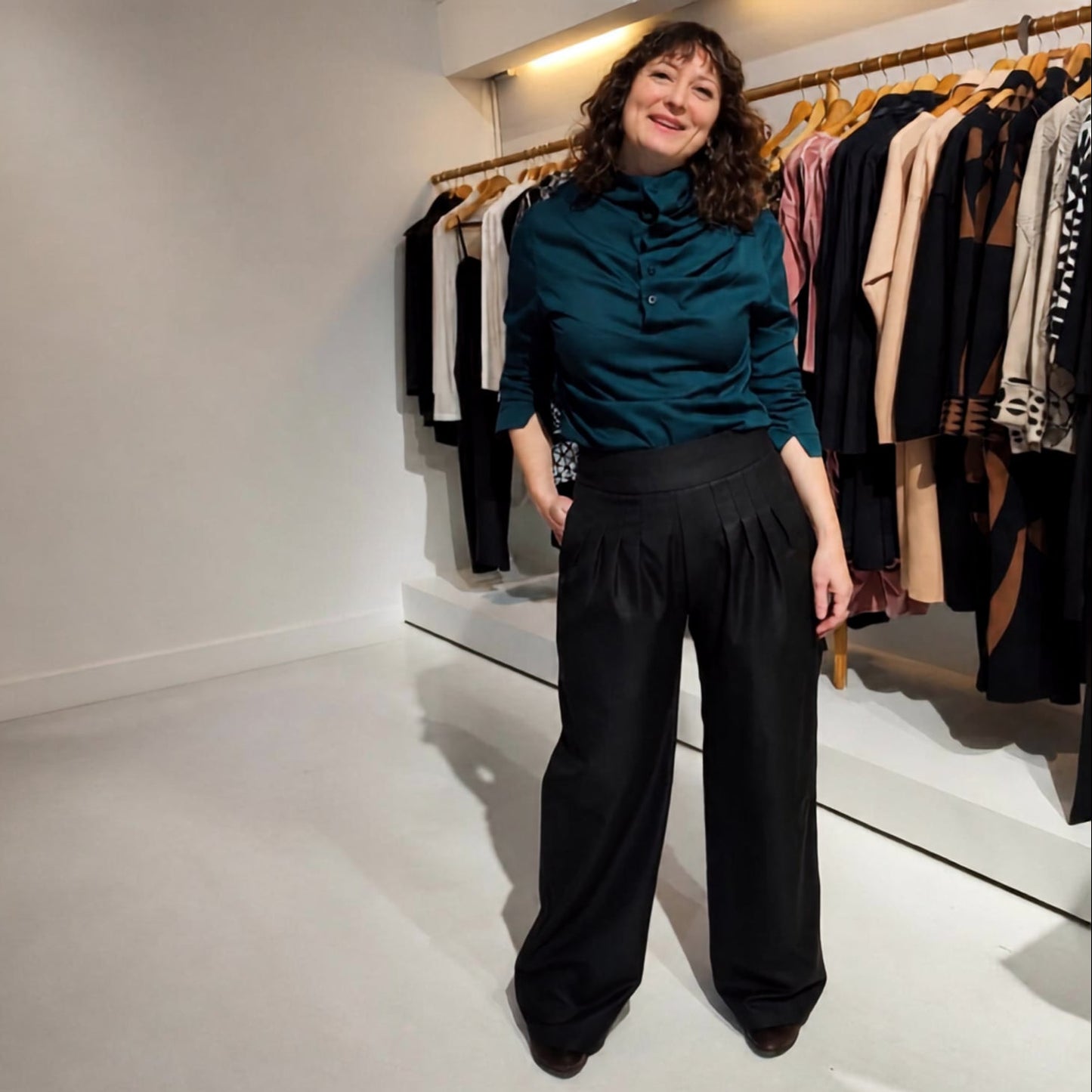 A woman stands in a boutique wearing a dark teal ruffled mock-neck blouse and black wide-leg trousers, with clothing racks visible in the background.