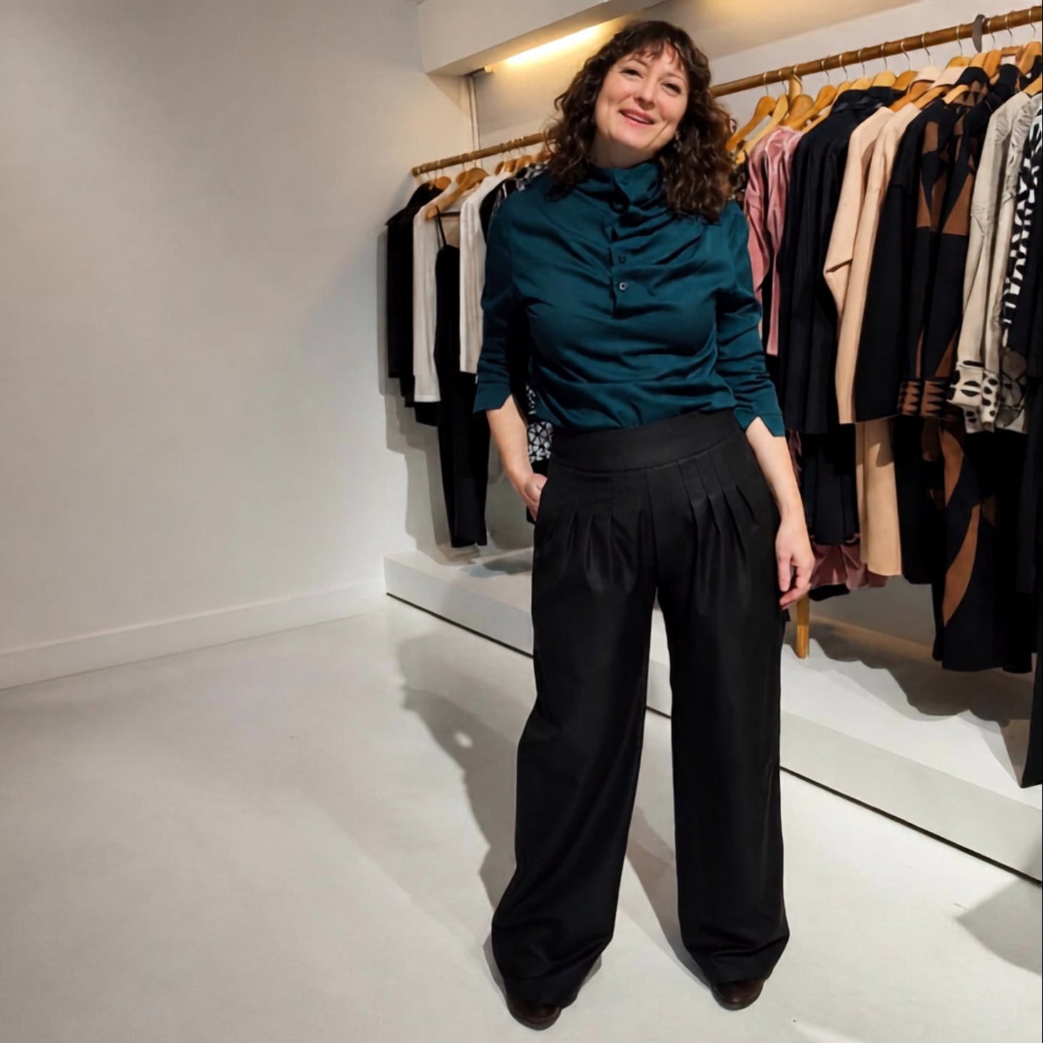 A woman stands in a boutique wearing a dark teal ruffled mock-neck blouse and black wide-leg trousers, with clothing racks visible in the background.