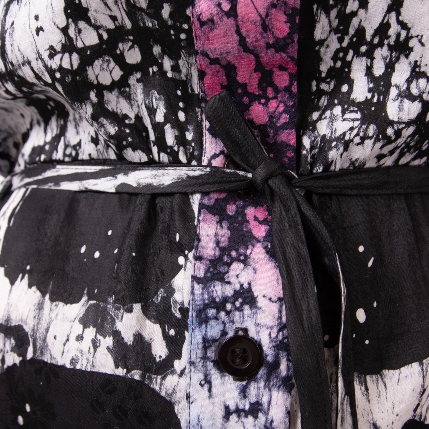 Close-up of a patterned garment with a black tie belt and dark button, featuring black, white, and pink abstract splatter and paint-stroke prints.